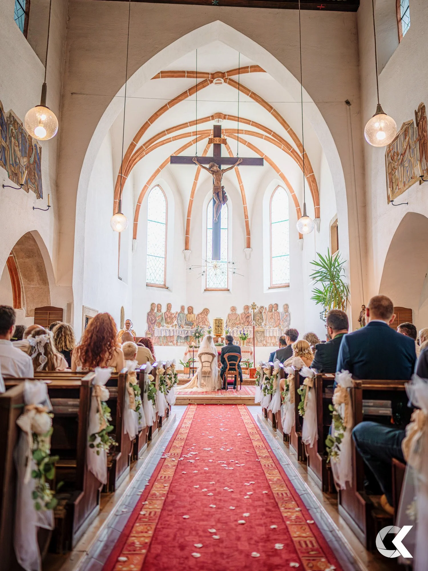 A wedding ceremony inside a church, with guests seated on wooden pews decorated with white ribbons and flowers, a red carpet leading to the altar, a bride and groom sitting at the front, a priest officiating, and a large crucifix hanging above the al