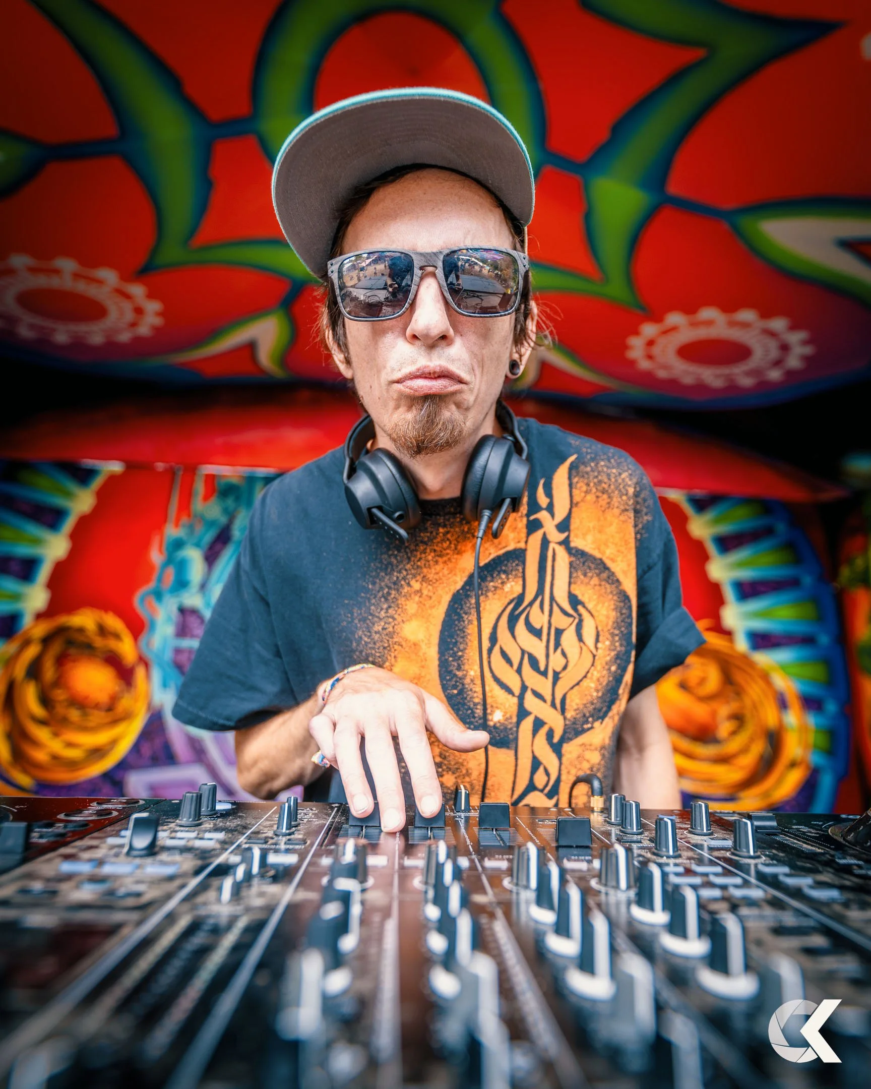A DJ wearing sunglasses and a cap adjusting controls on a mixing console with colorful psychedelic wall art in the background.