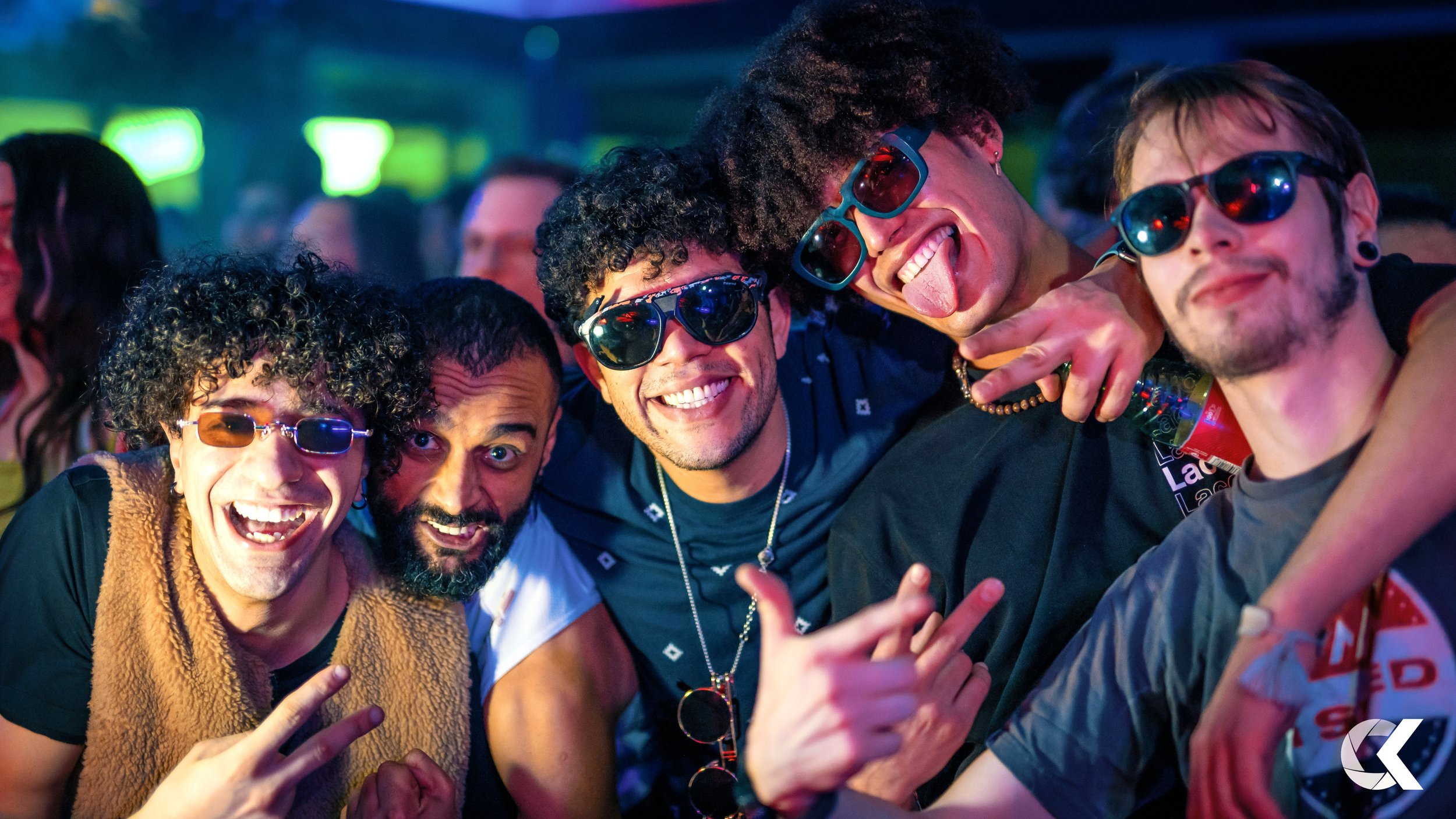 A group of five friends at a party or concert, smiling and wearing sunglasses, with colorful lights in the background.