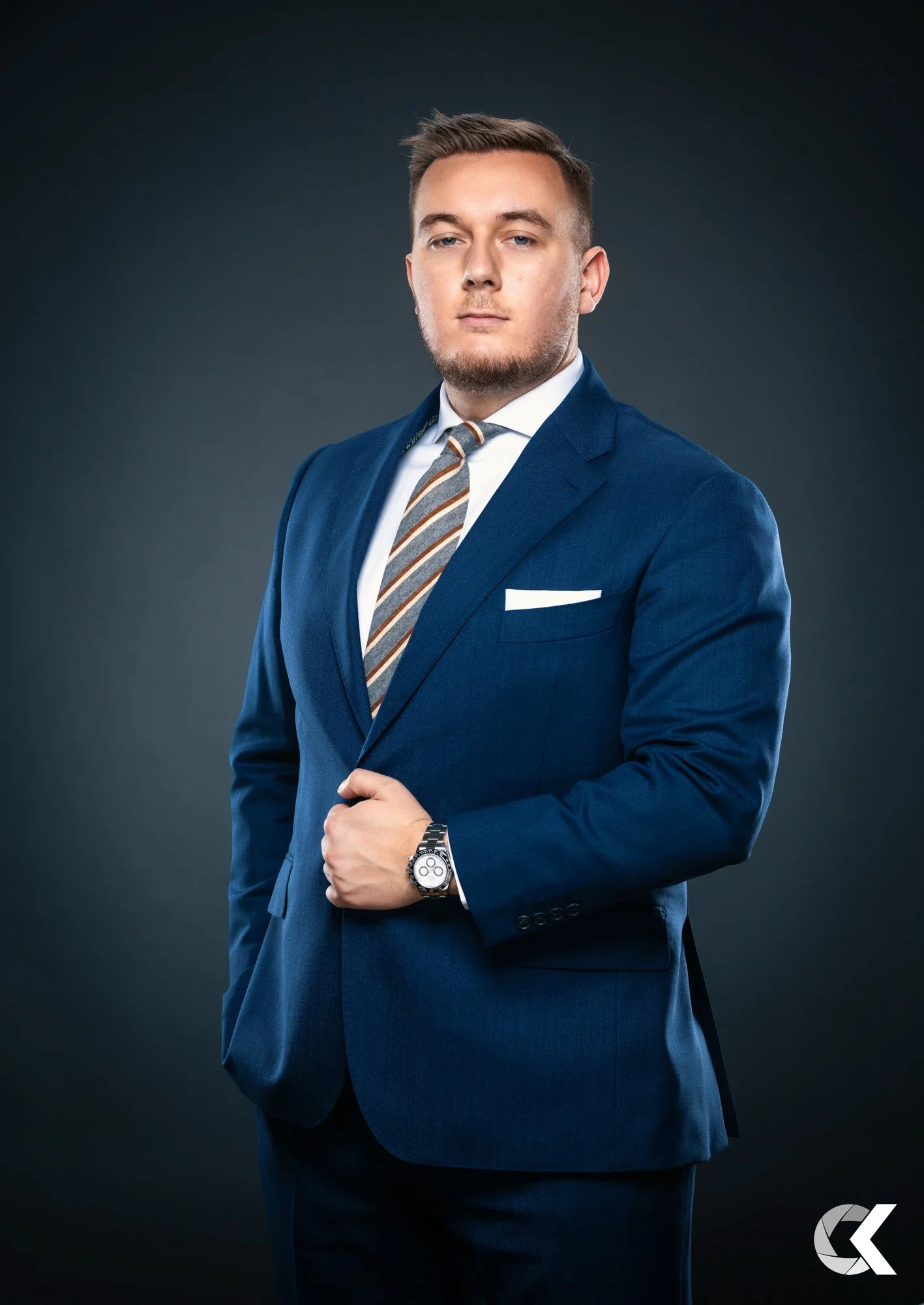 A young man in a blue suit with a striped tie, white shirt, and a watch, posing against a dark background.
