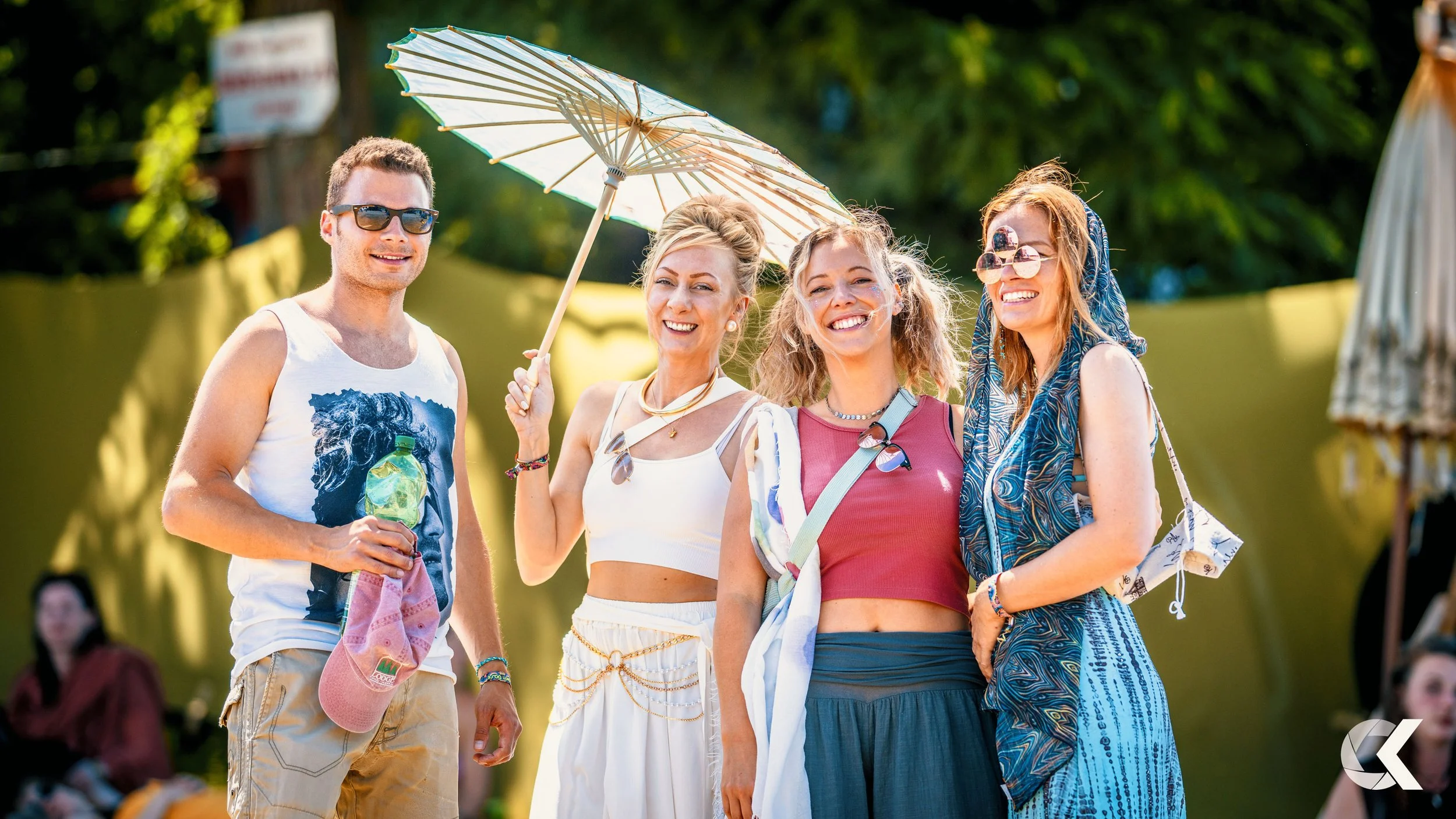 Four young adults at an outdoor event on a sunny day, smiling, with two women holding a parasol, and wearing casual summer clothes.