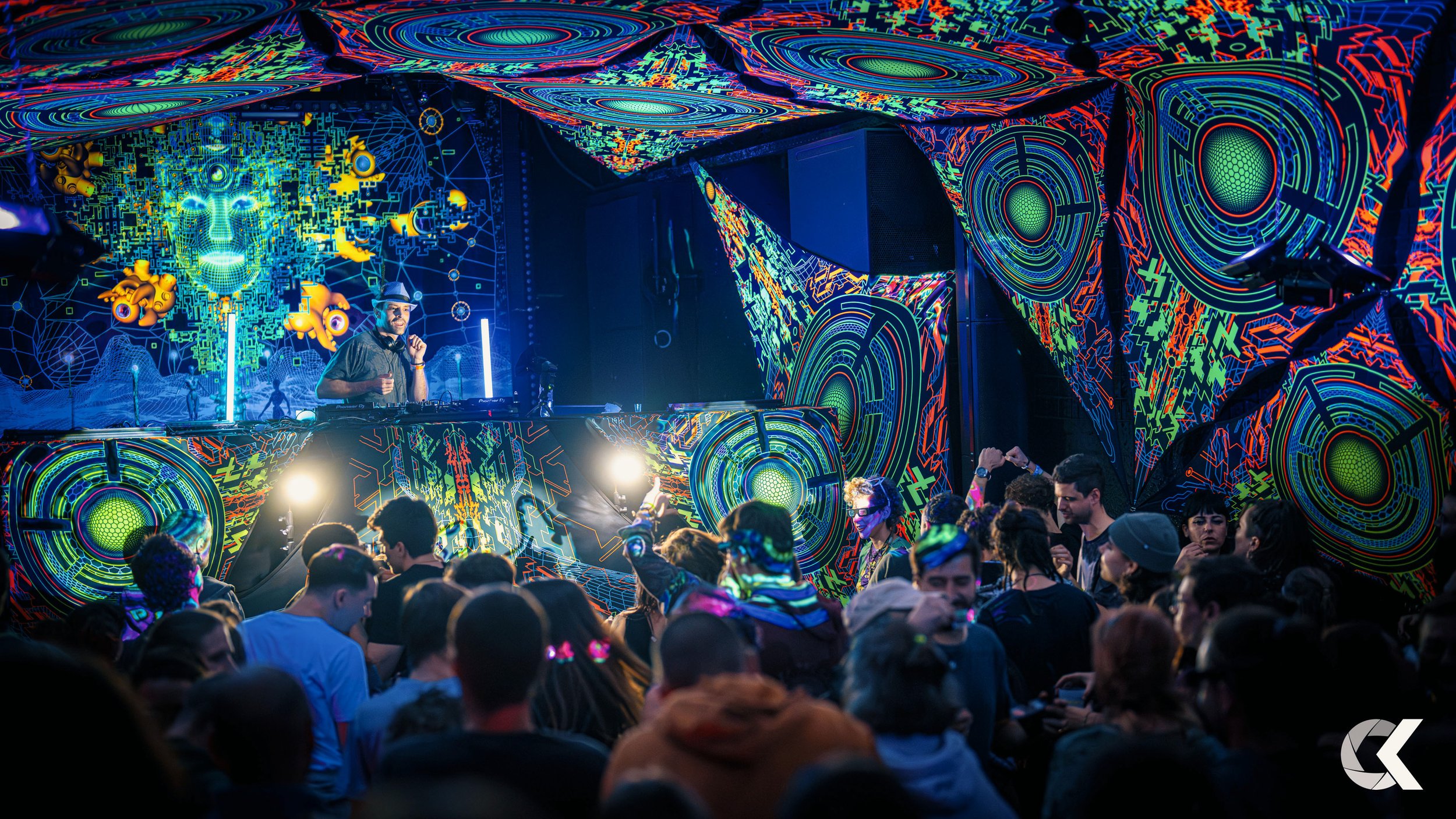 A vibrant electronic music concert with a DJ performing on stage, decorated with colorful digital and geometric designs. The crowd is dancing and enjoying the event, with some people wearing glow-in-the-dark accessories.