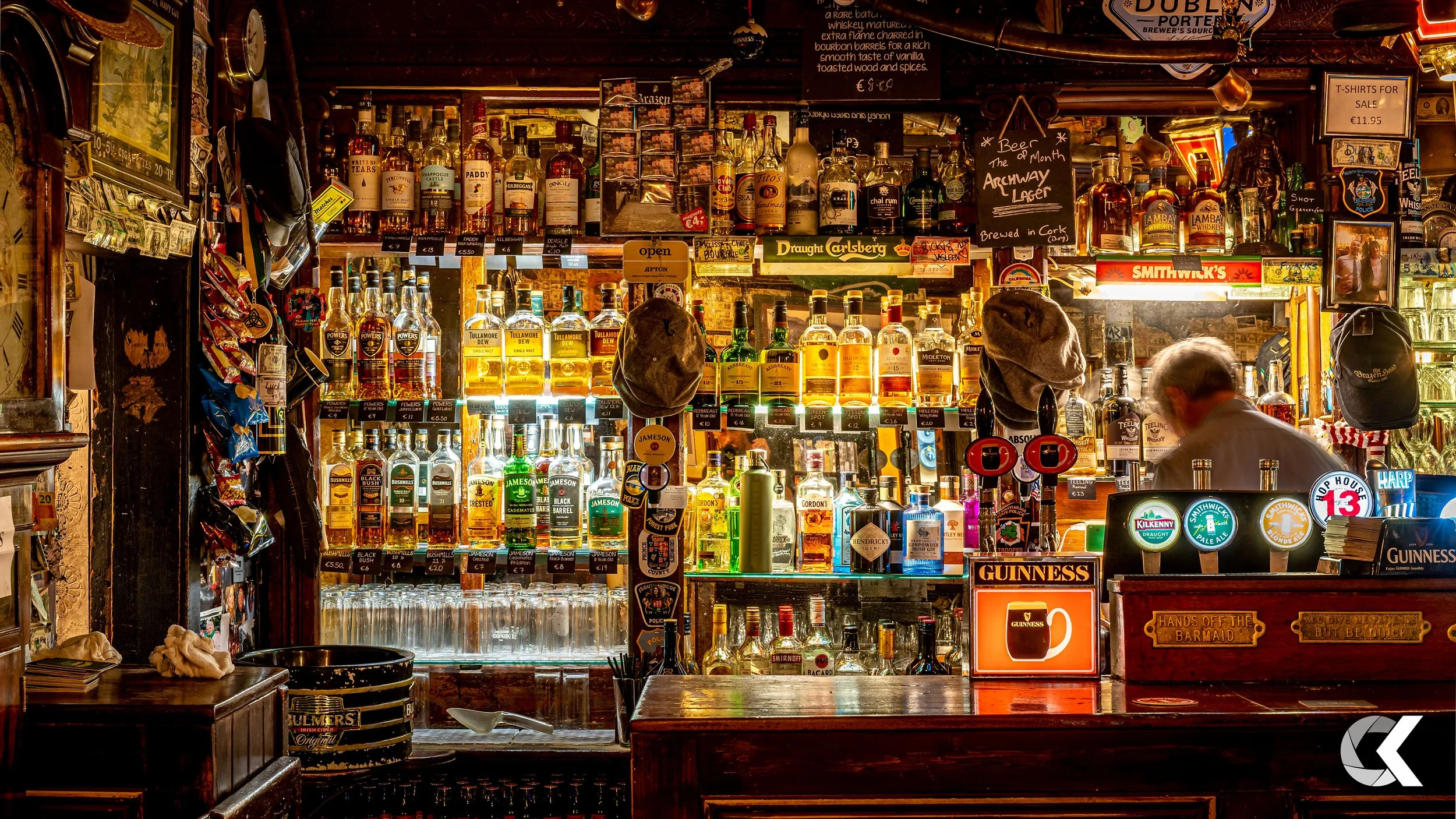 A bar with shelves filled with various bottles of alcohol, including whiskey, gin, and beer. The bar has illuminated signs for Guinness and Jameson, hats hanging on display, and a customer with gray hair leaning on the bar counter. There are signs an