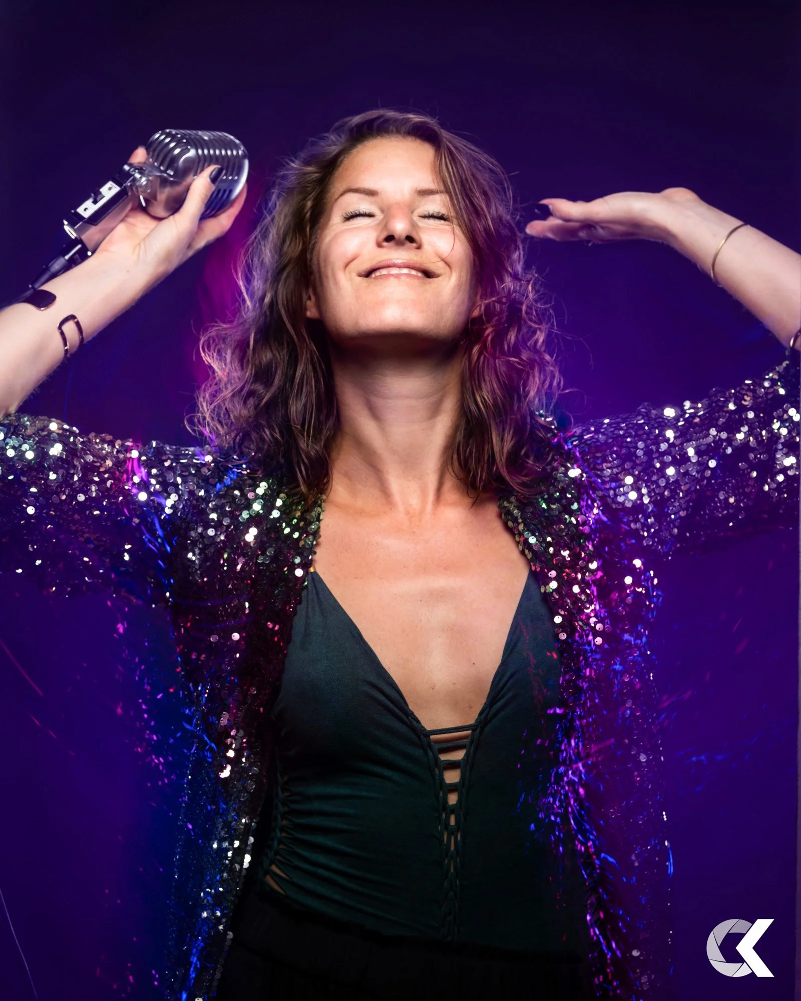A woman with shoulder-length wavy hair is smiling with eyes closed, holding a vintage microphone in her left hand, and raising her right hand near her head. She is wearing a sparkling, sequined jacket over a black top with lace-up details.