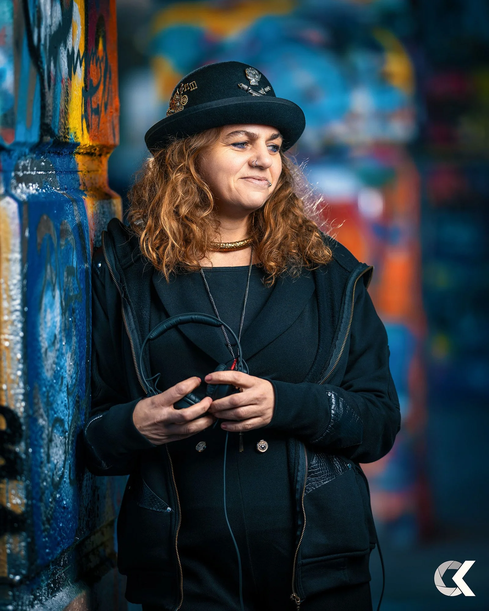 A young woman with curly red hair wearing a black hat with pins, a black hoodie, and a gold choker, holding headphones while leaning against a colorful graffiti-covered wall.