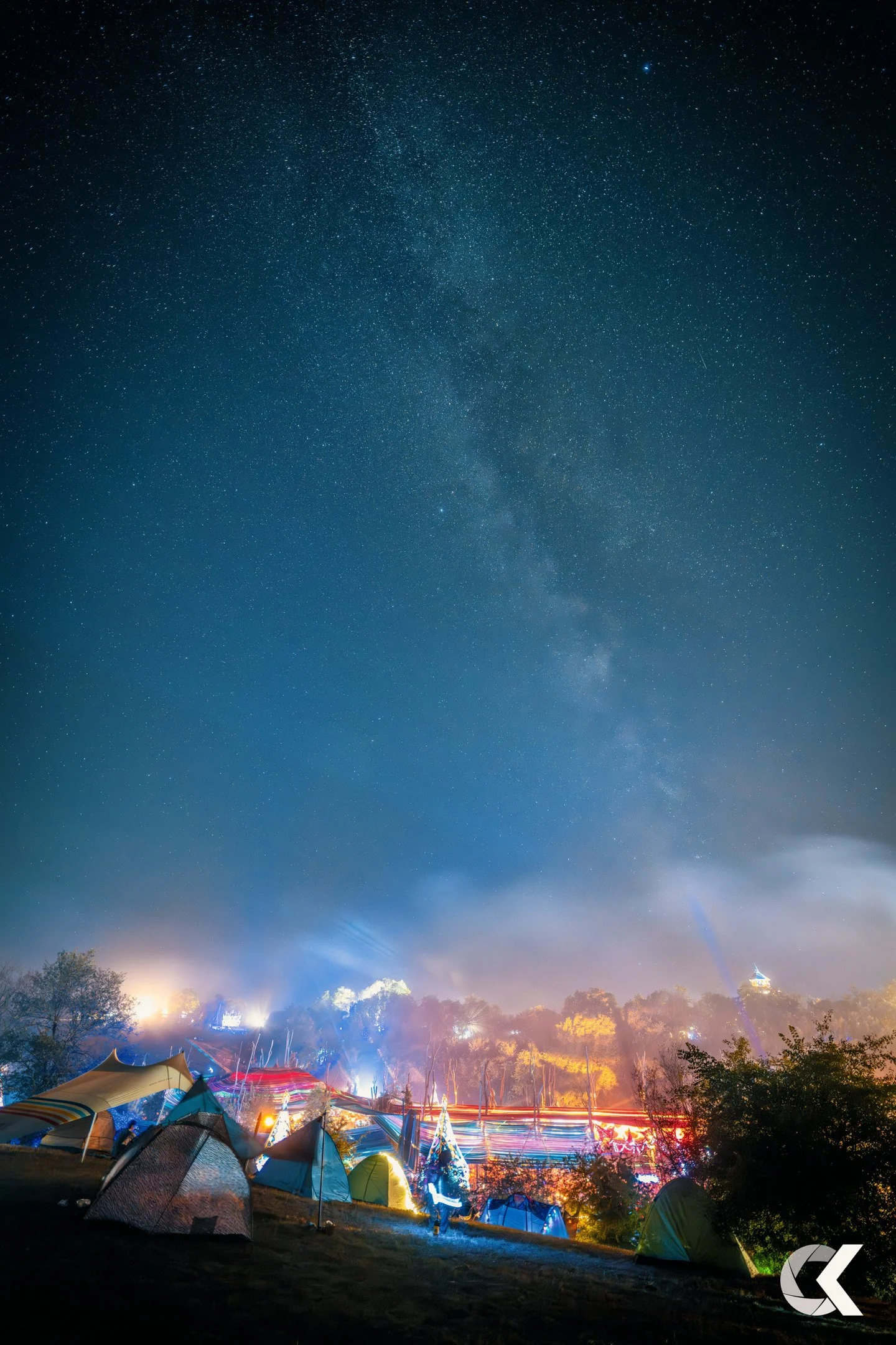 Nighttime scene at a camping area with tents illuminated near trees, bright lights, and a clear starry sky featuring the Milky Way galaxy.