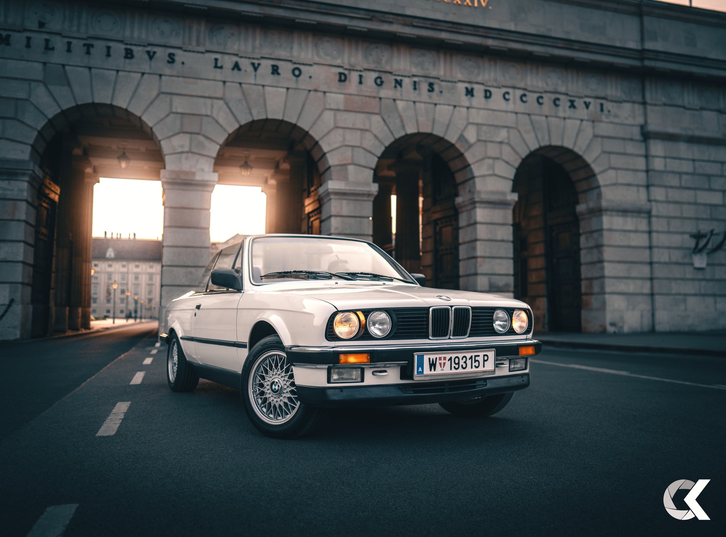 A classic white BMW car parked on a city street in front of a stone building with arched openings.