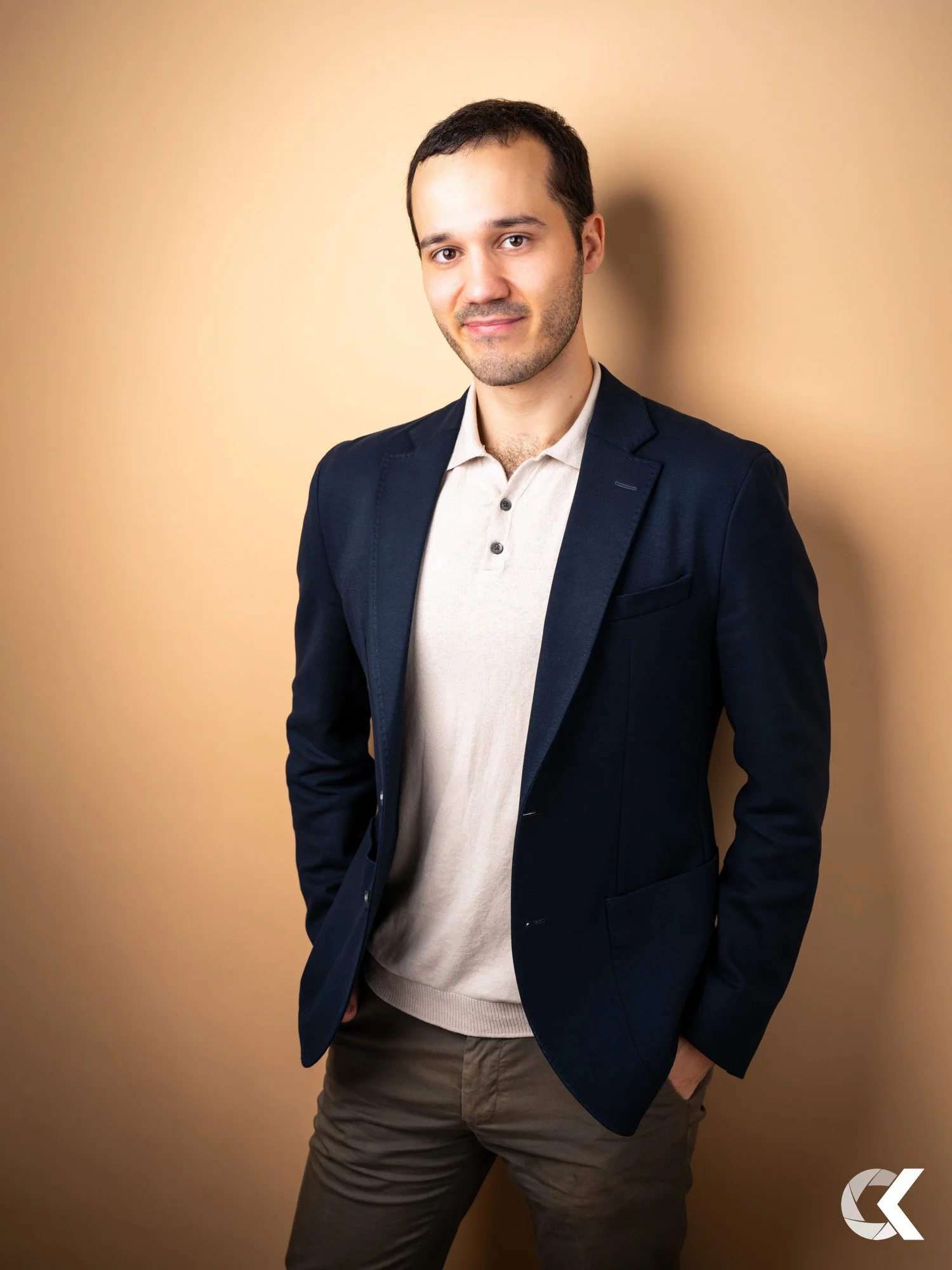 A man in a navy blazer and light-colored polo shirt poses against a plain beige background, with his hands in his pockets and a slight smile.