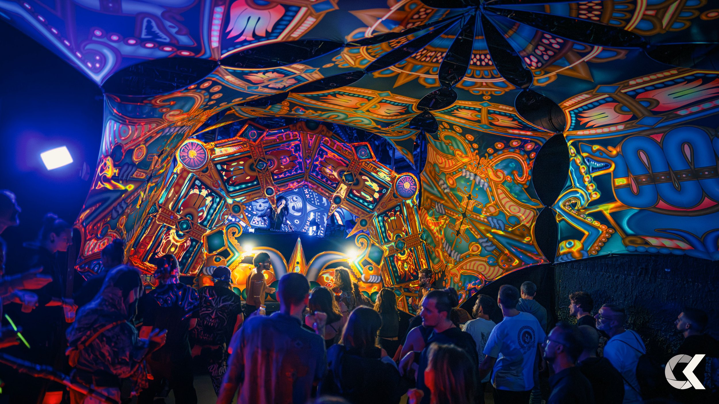 Crowd of people at a music festival with a colorful, psychedelic stage backdrop featuring intricate designs and vibrant colors.