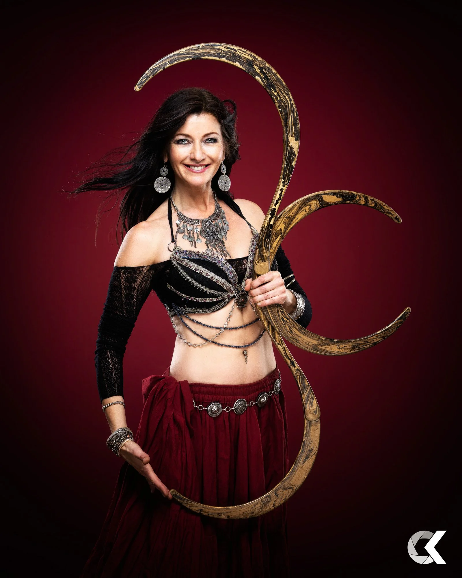 A woman with long dark hair wearing bohemian jewelry and a black lace crop top holding a large decorative crescent-shaped metal staff, standing against a dark red gradient background.