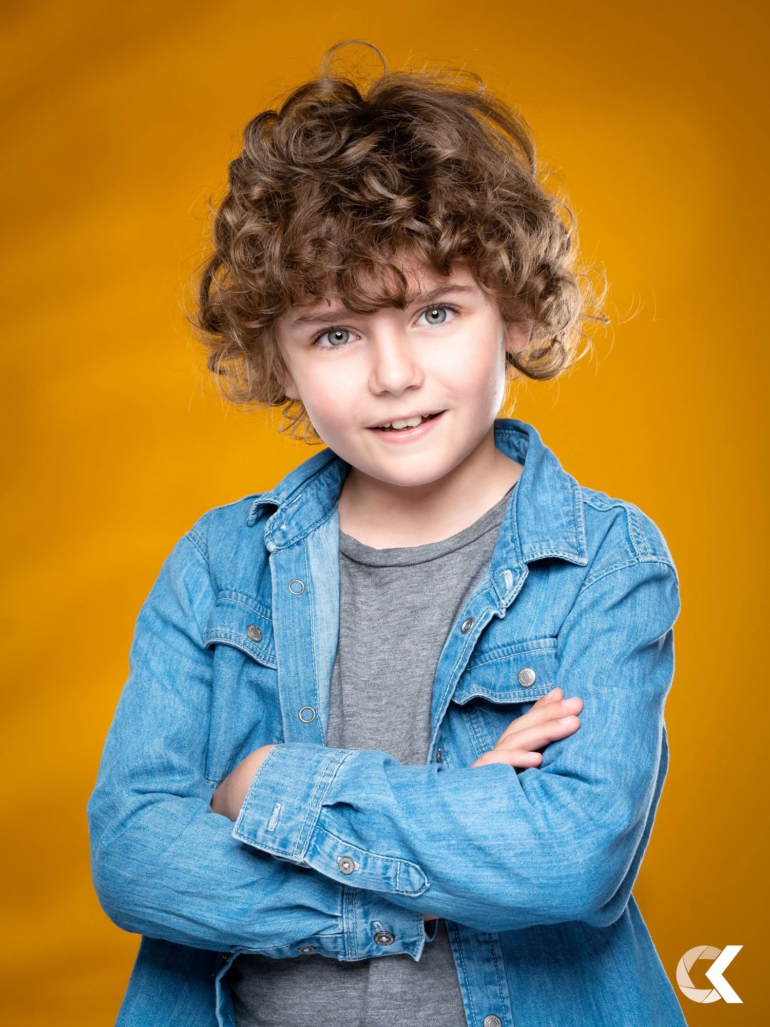 A young boy with curly hair and blue eyes is standing against a yellow background, smiling and wearing a denim jacket over a gray shirt.