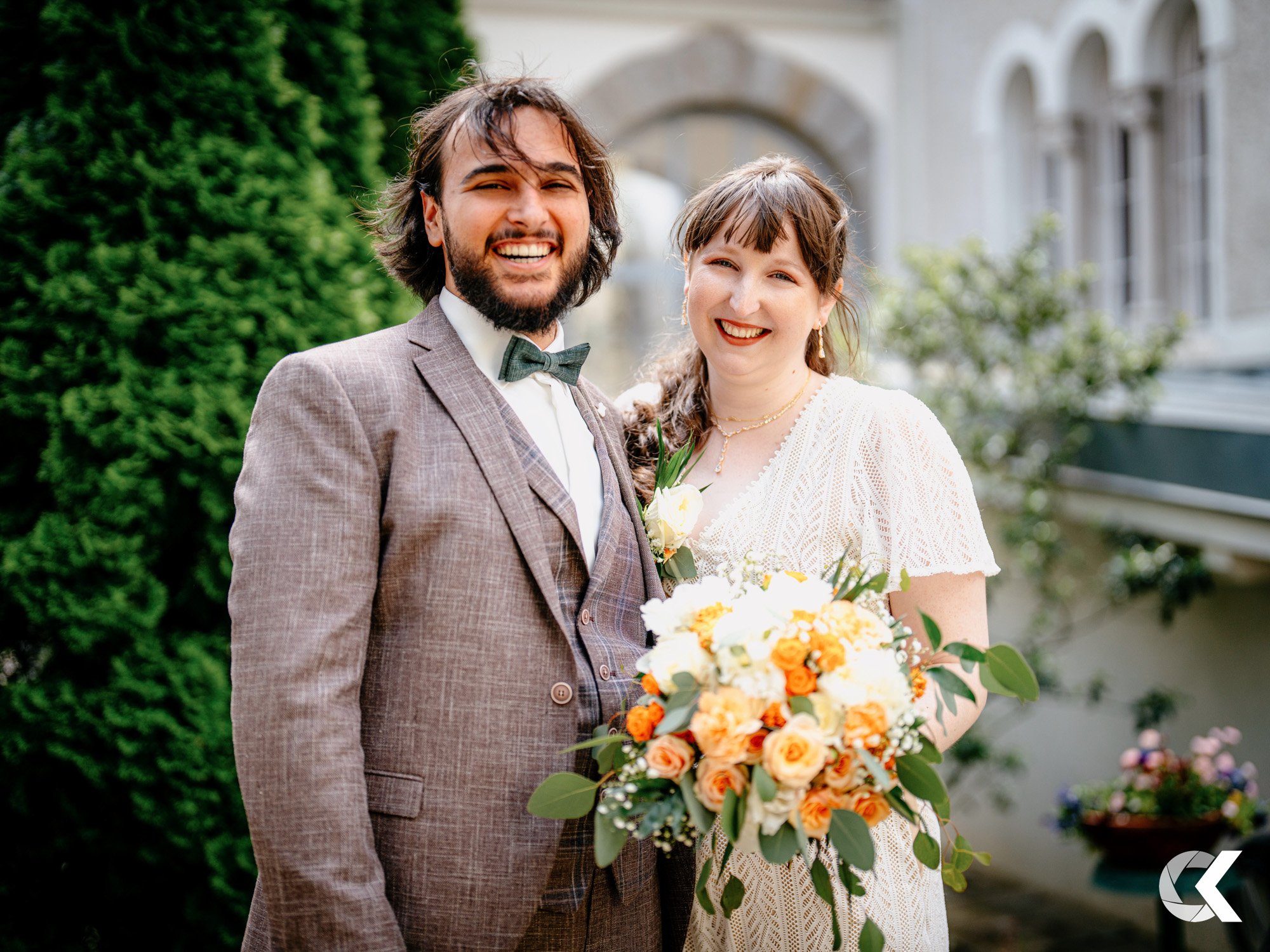 A happy bride and groom outdoors, smiling at the camera. The groom wears a plaid suit with a bowtie, and the bride holds a bouquet of orange, white, and green flowers, dressed in a white lace dress.