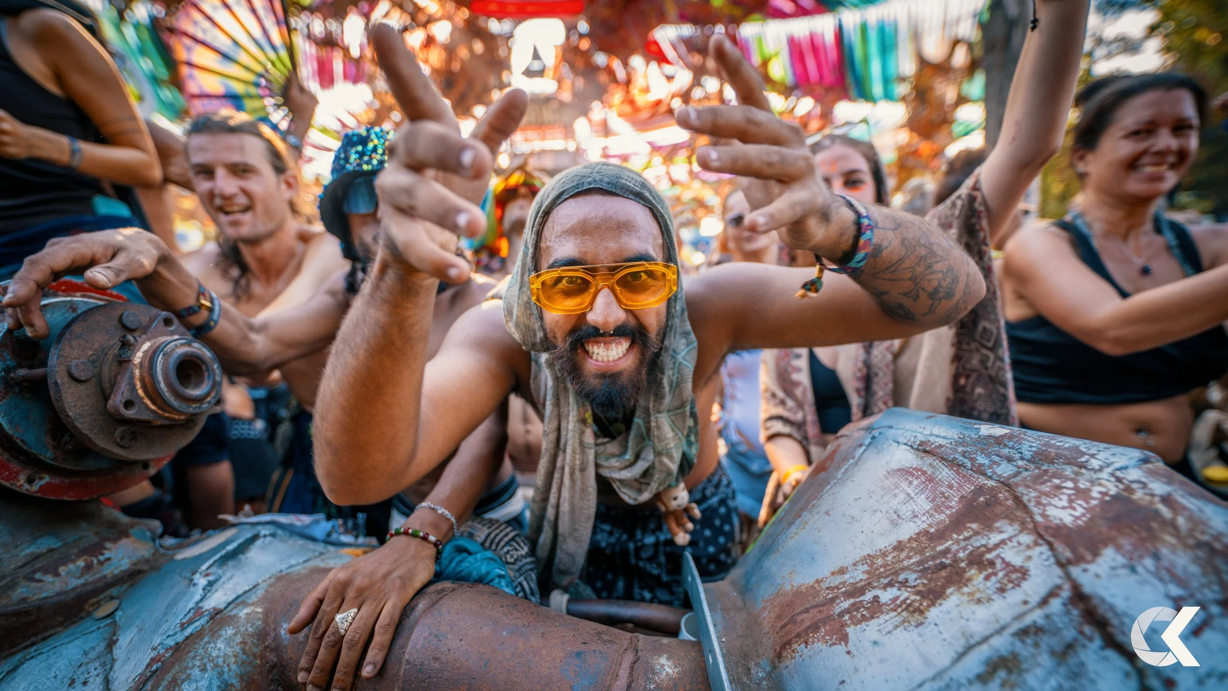 A group of people at a festival or event, with a bearded man wearing yellow-tinted sunglasses in the center, smiling and making a peace sign, surrounded by colorful decorations and other smiling attendees.