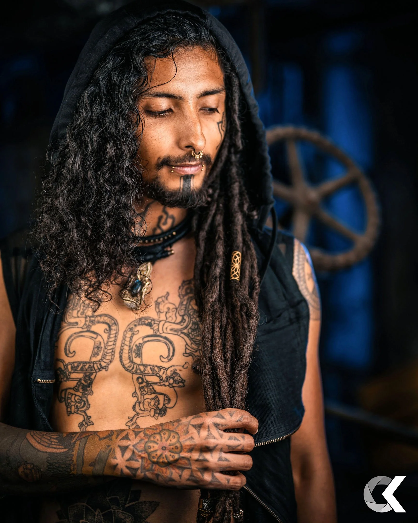 A man with long curly hair, dreadlocks, and tattoos on his chest and arm, wearing a black hoodie and jewelry, looking down with a thoughtful expression in a dimly lit environment.