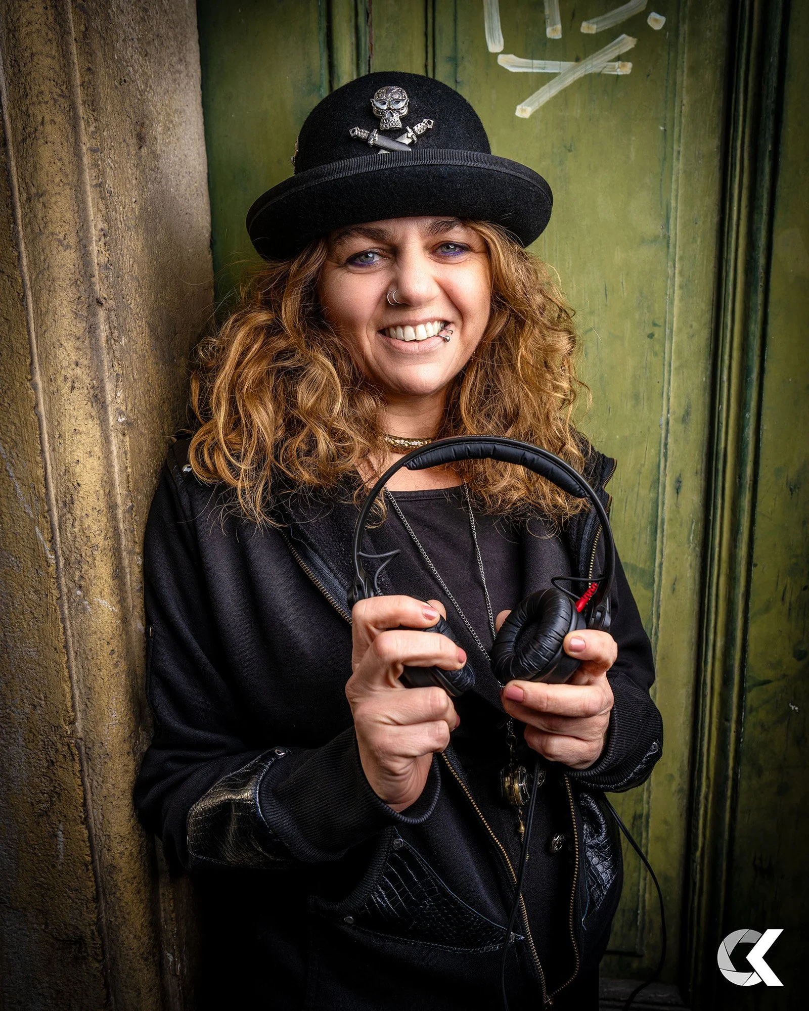 A woman with curly hair and piercings smiling, wearing a black hat with a skull and crossbones pin, holding headphones in her hands, standing against a weathered green door and stone wall background.