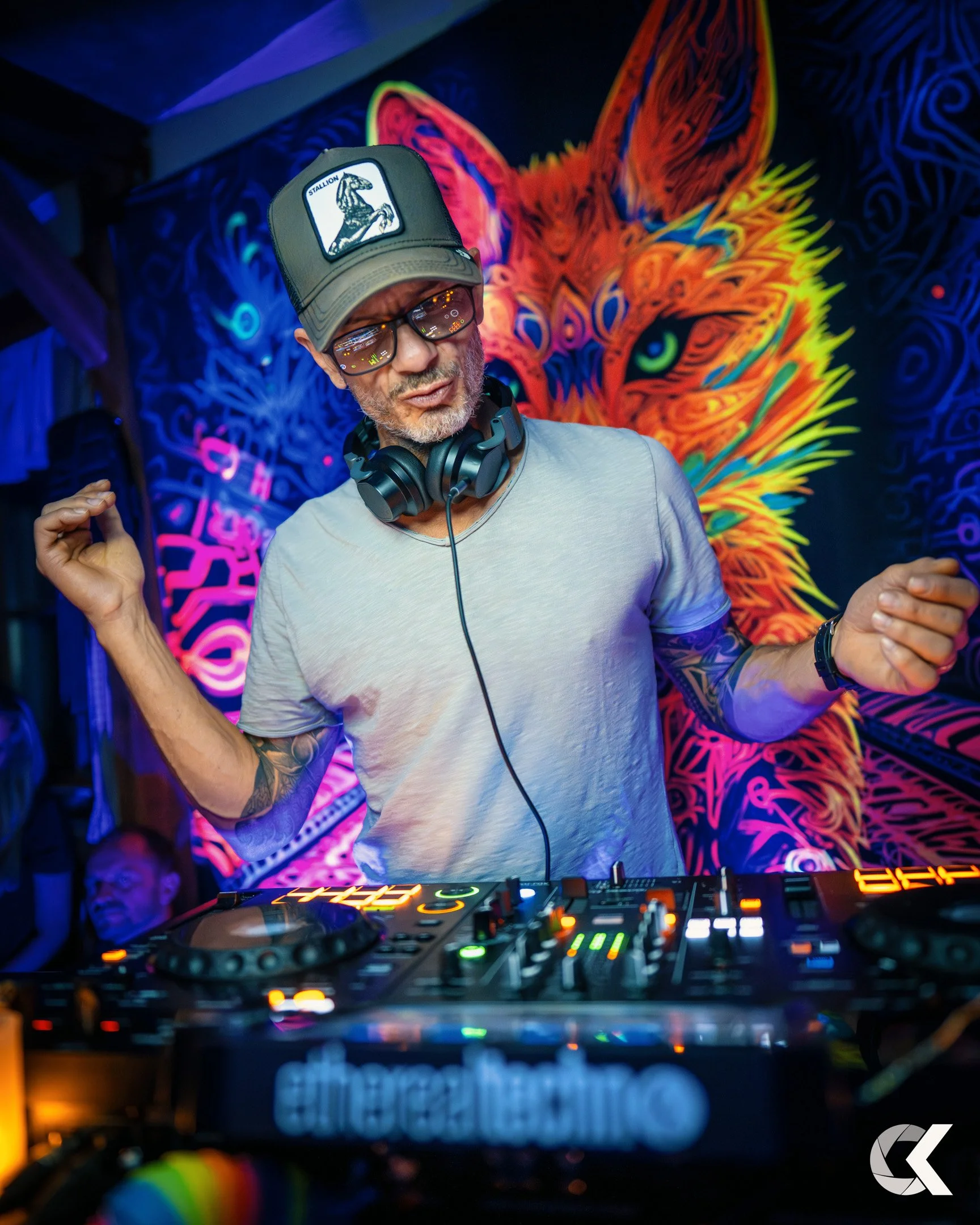 A DJ with headphones around his neck, wearing a hat and glasses, dancing in front of a colorful tiger mural at a nightclub or party.