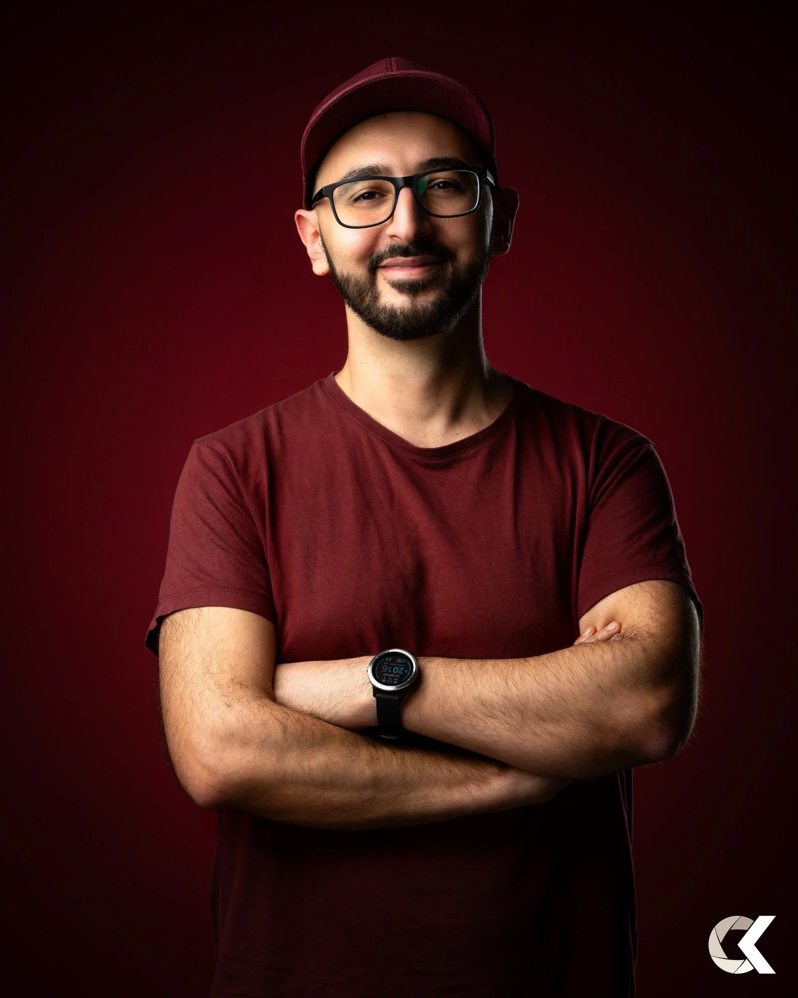 A man wearing a maroon t-shirt, maroon cap, glasses, and a smartwatch with arms crossed. He is smiling against a dark red background.