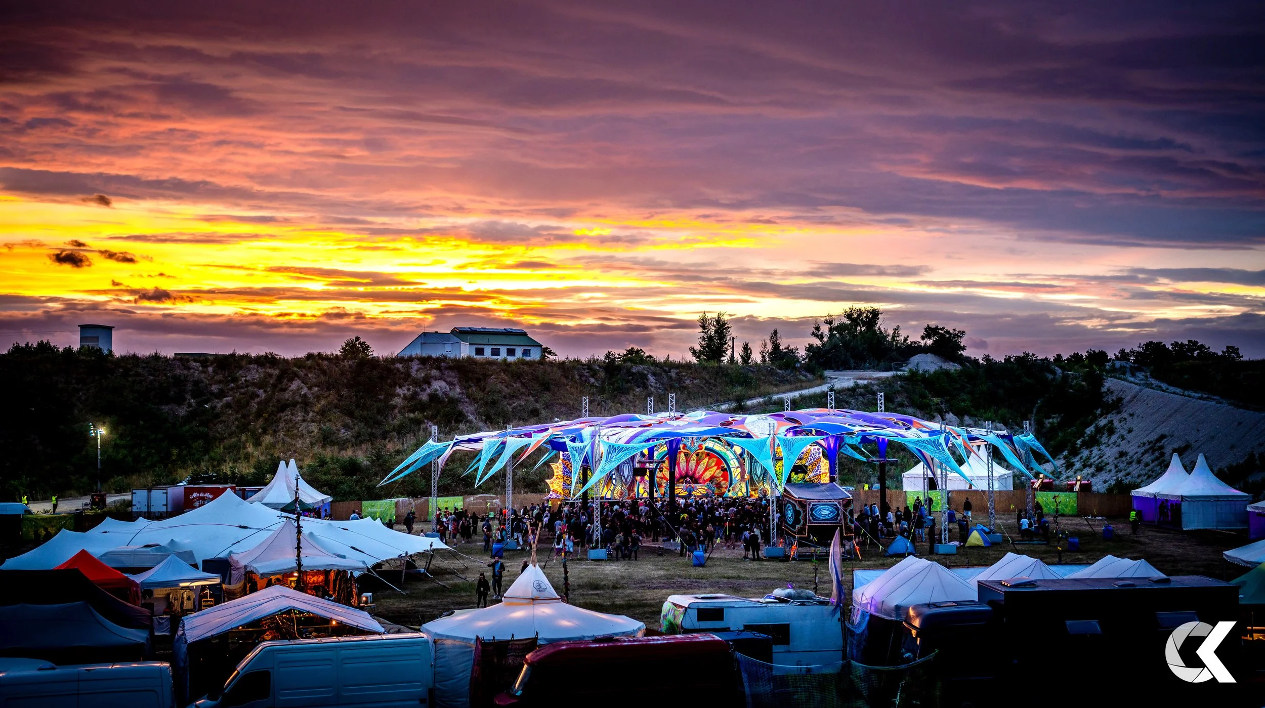 Sunset over a music festival with a colorful stage, tents, and crowds of people.