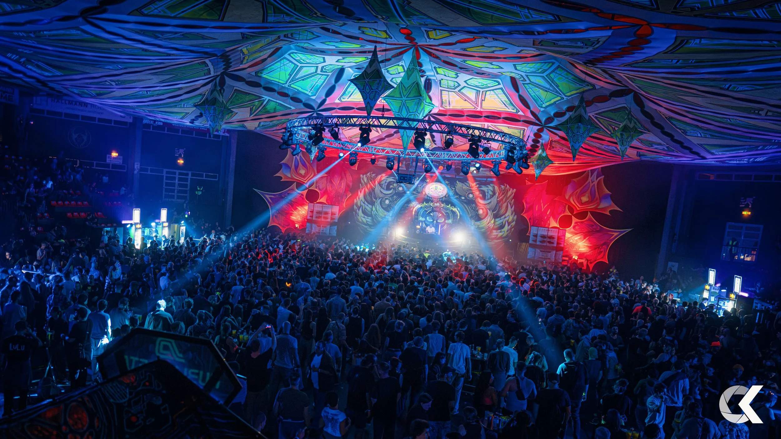 Crowd of people at a concert or music event with colorful lighting, illuminated screens, and artistic hanging decorations on the ceiling.