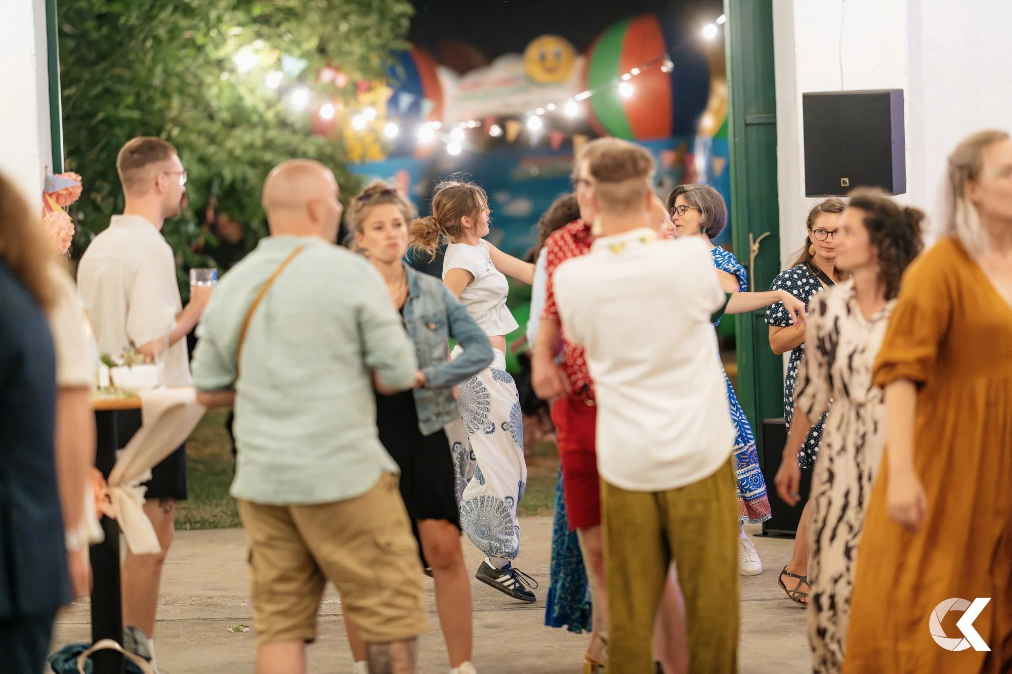 People dancing and socializing at an outdoor party or event during evening, with colorful decorations and string lights in the background.