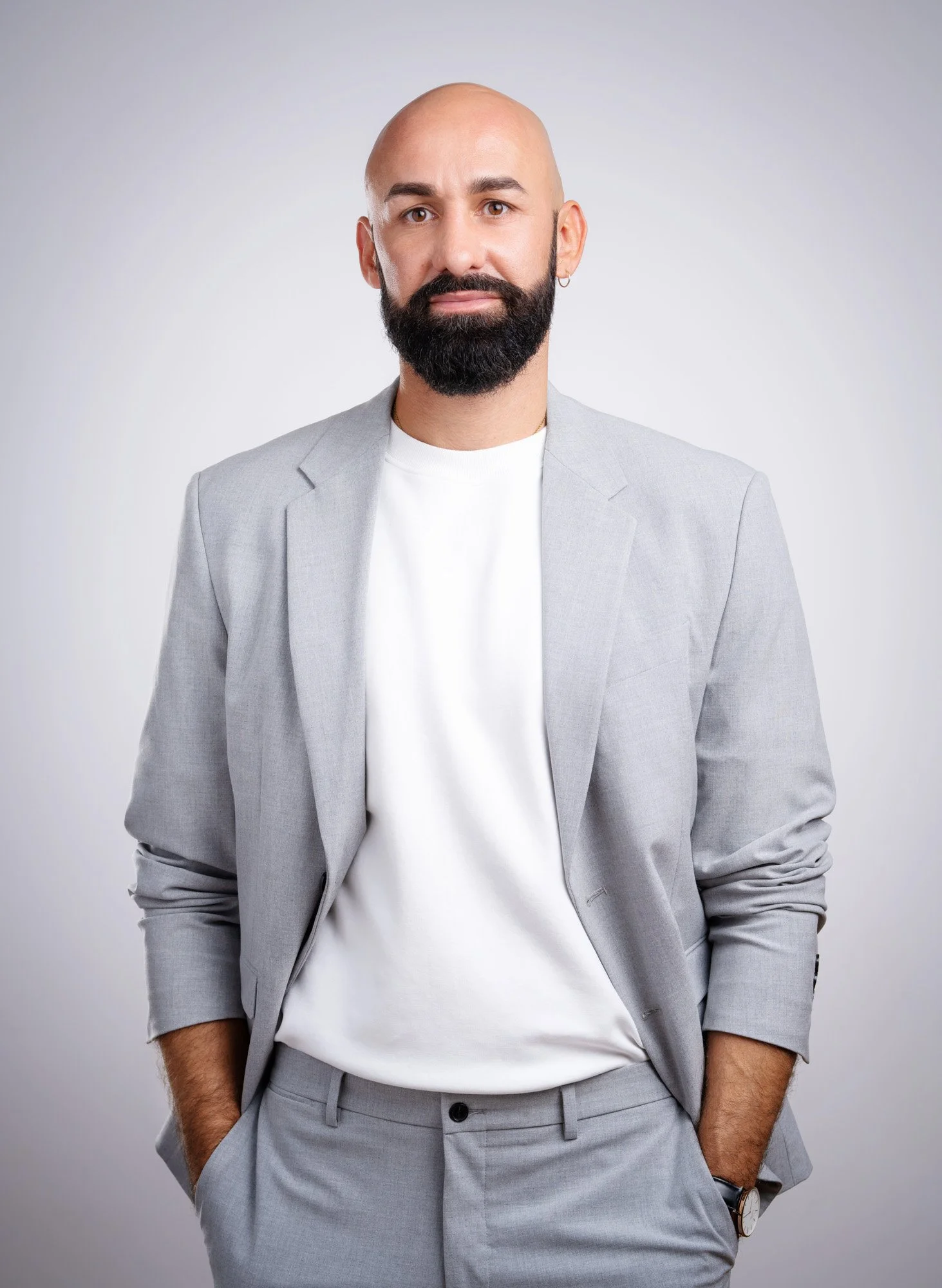 A bald man with a beard and earring wearing a light gray suit with a white T-shirt inside, standing against a gray background.