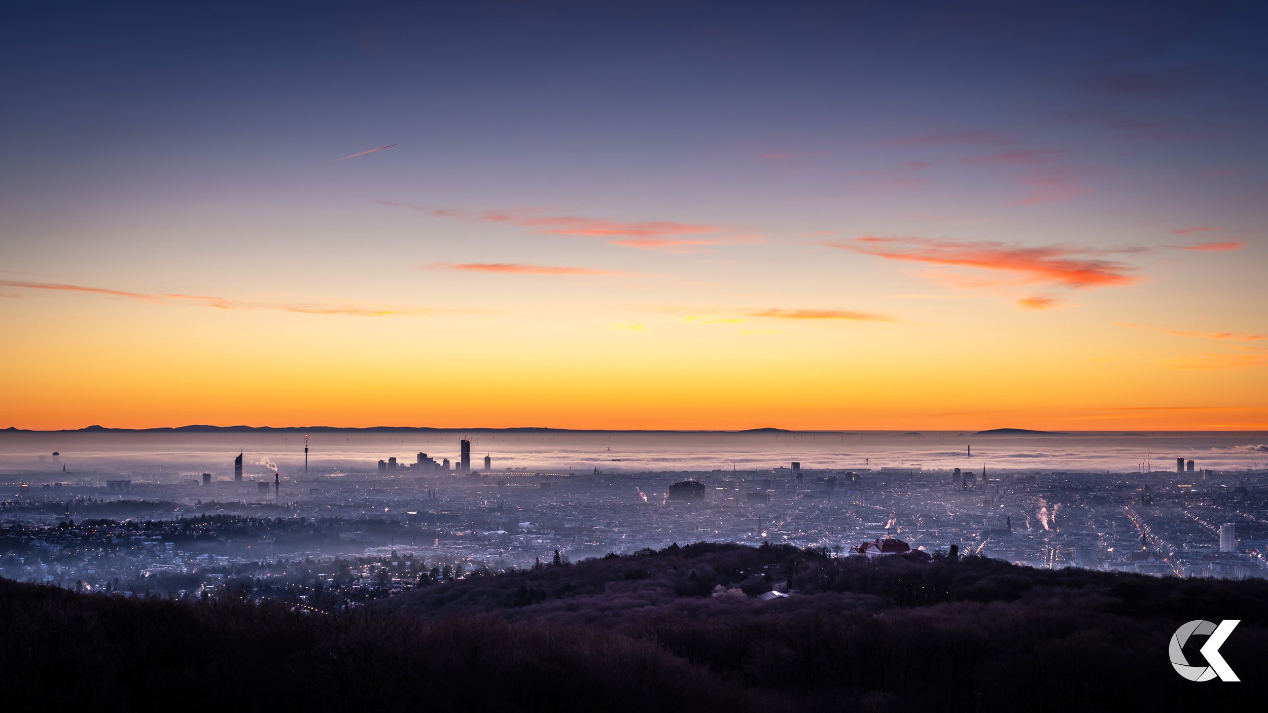 Sunrise over a city skyline with tall buildings, smokestacks, and a layer of fog or mist in the foreground.