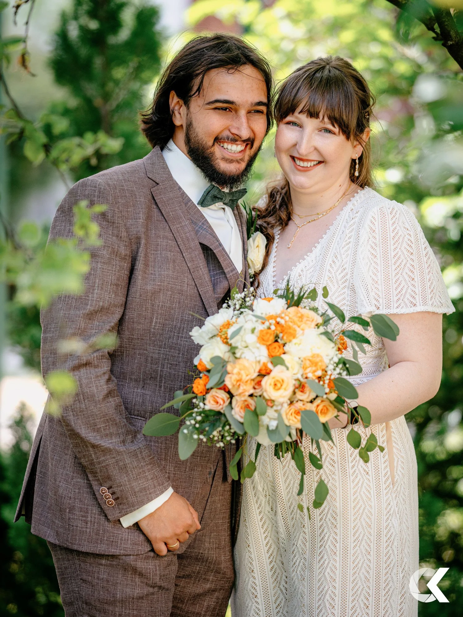 A happy couple dressed in wedding attire, standing outdoors with greenery in the background. The man is wearing a brown suit with a green bow tie, and the woman is in a white dress with lace details, holding a bouquet of white, orange, and yellow flo