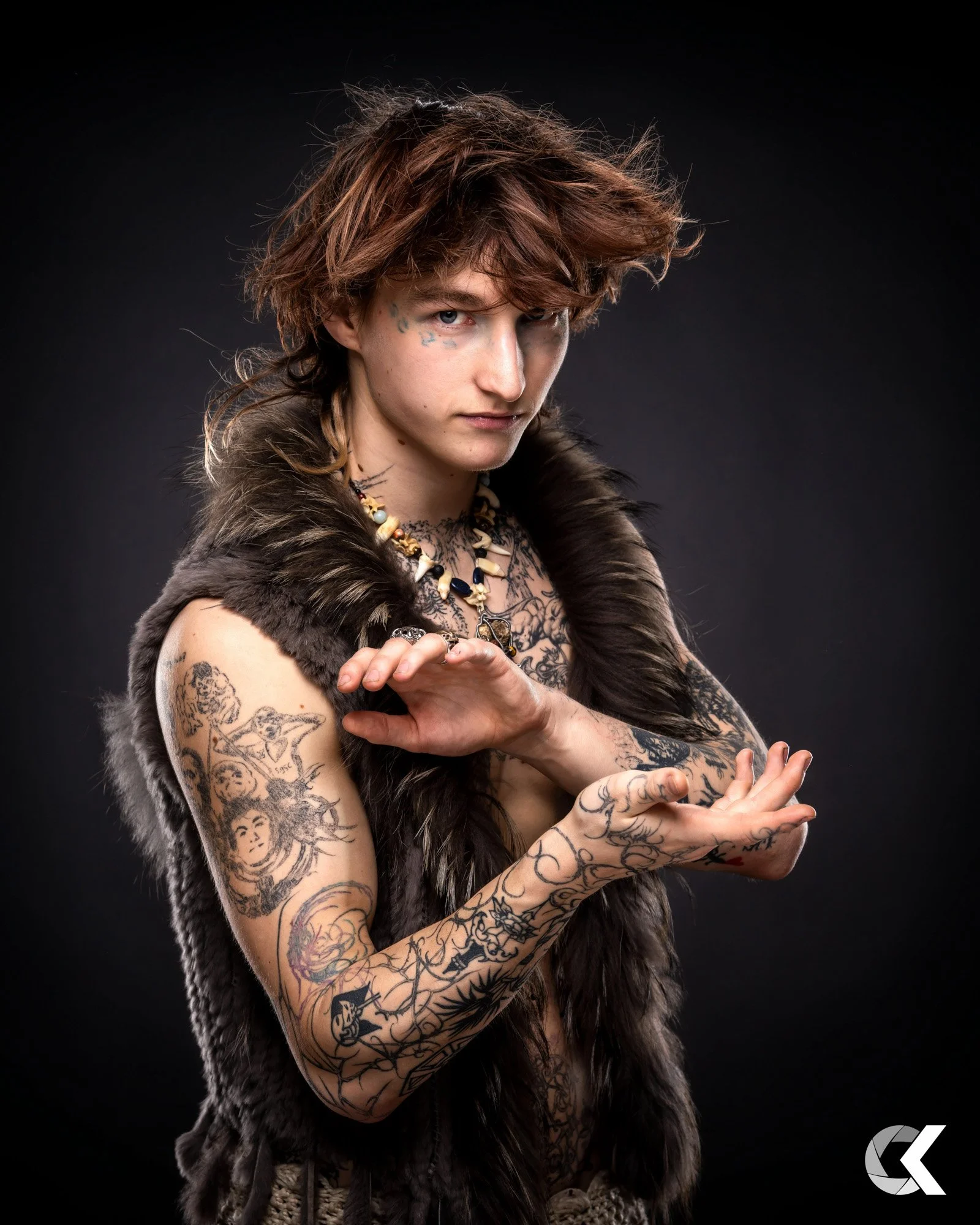 Portrait of a tattooed young person with brown, tousled hair, wearing a fur vest, and jewelry, posing with hands raised against a dark background.