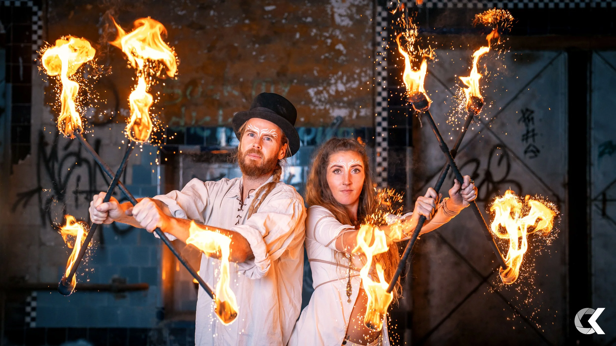 Two performers in vintage style clothing and makeup hold flaming torches in a theatrical performance, with graffiti and industrial background.