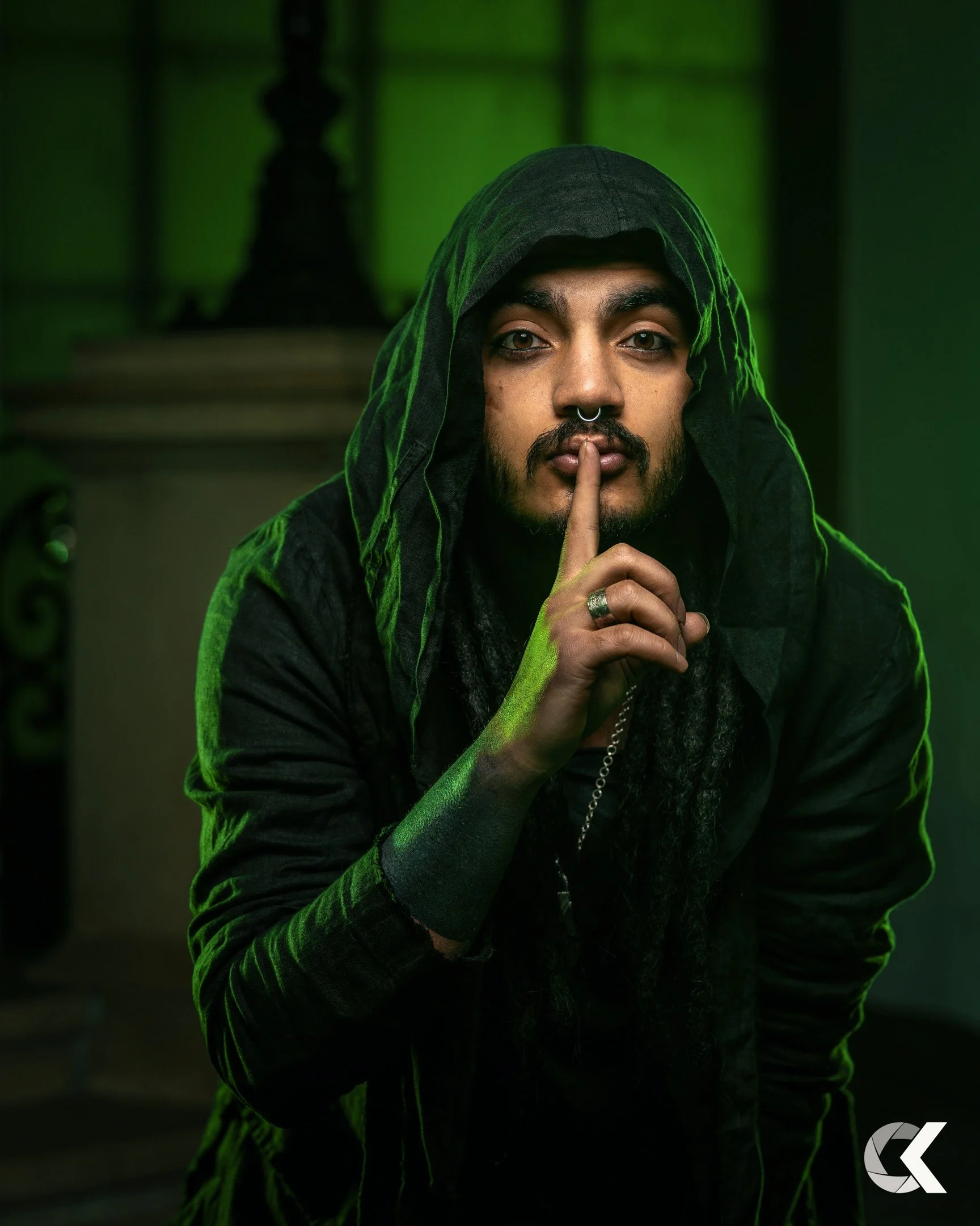 Person with long dreadlocks, wearing a hooded jacket, holding a finger to their lips in a shushing gesture, in a green-lit room.