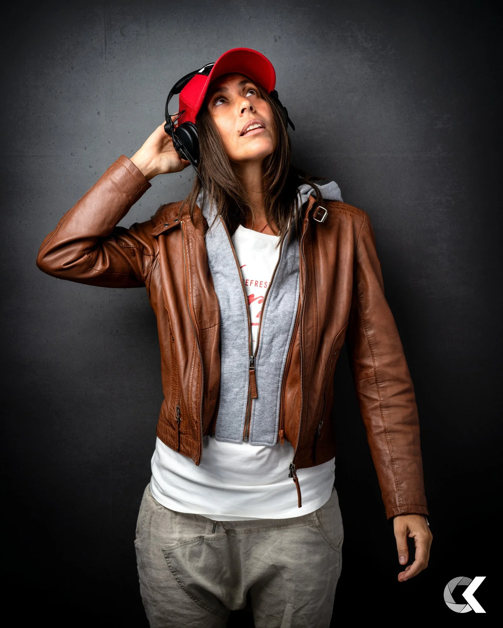 Woman wearing a red cap and brown leather jacket puts on headphones while looking up, standing against a dark gray background.