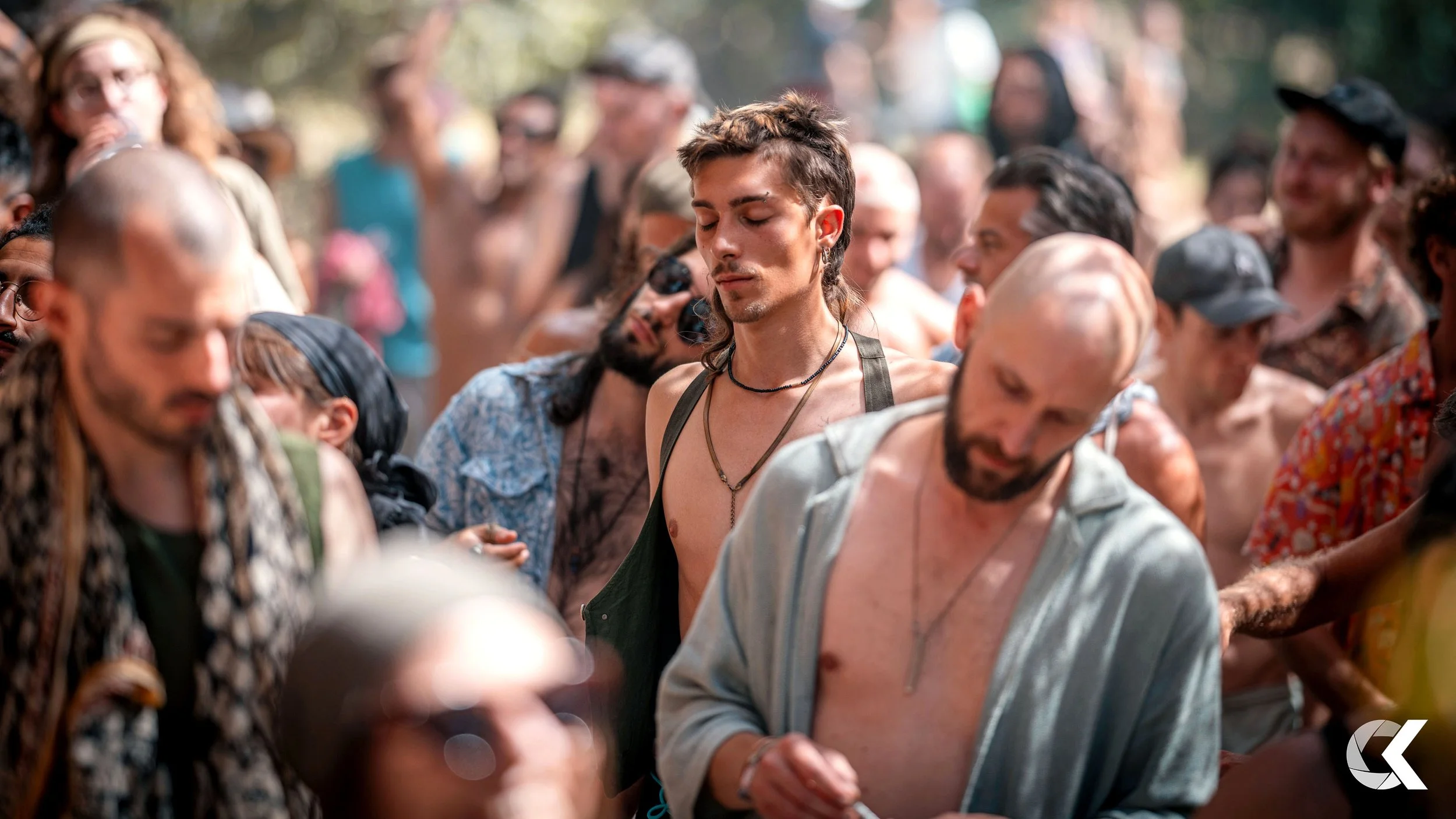 A group of people gathered outdoors, with many appearing to have their eyes closed or looking down, and some have their shirts off or are shirtless. The scene suggests a communal or spiritual gathering.