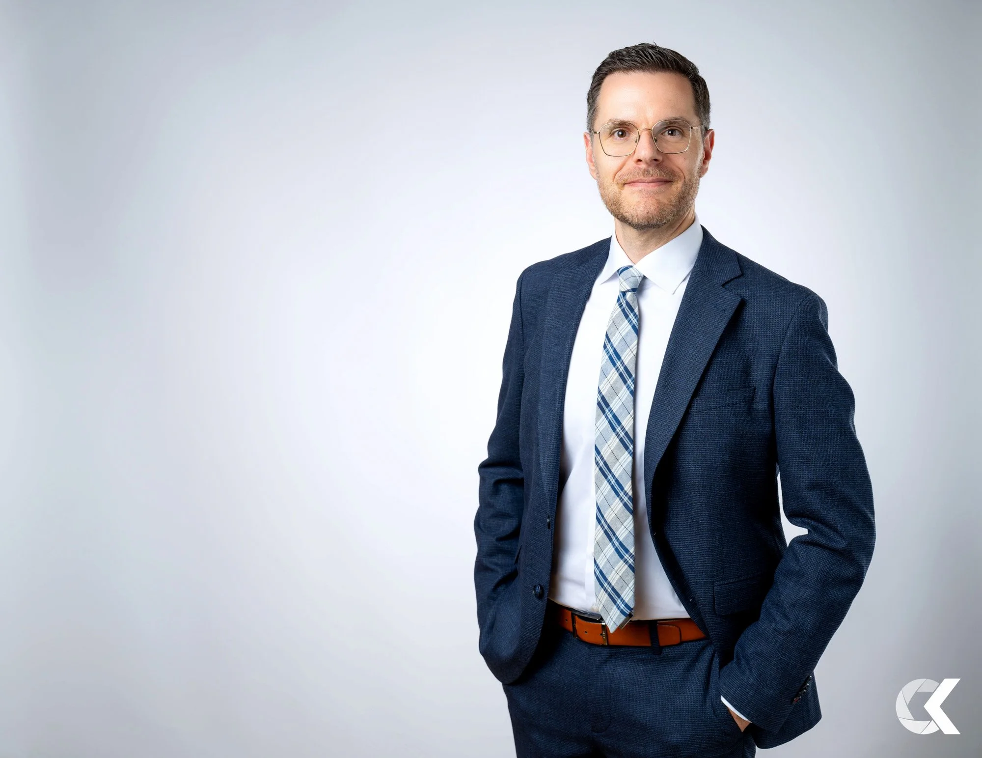 A man in a dark blue suit, white dress shirt, and plaid tie standing with his right hand in his pocket, smiling slightly, against a plain gray background.