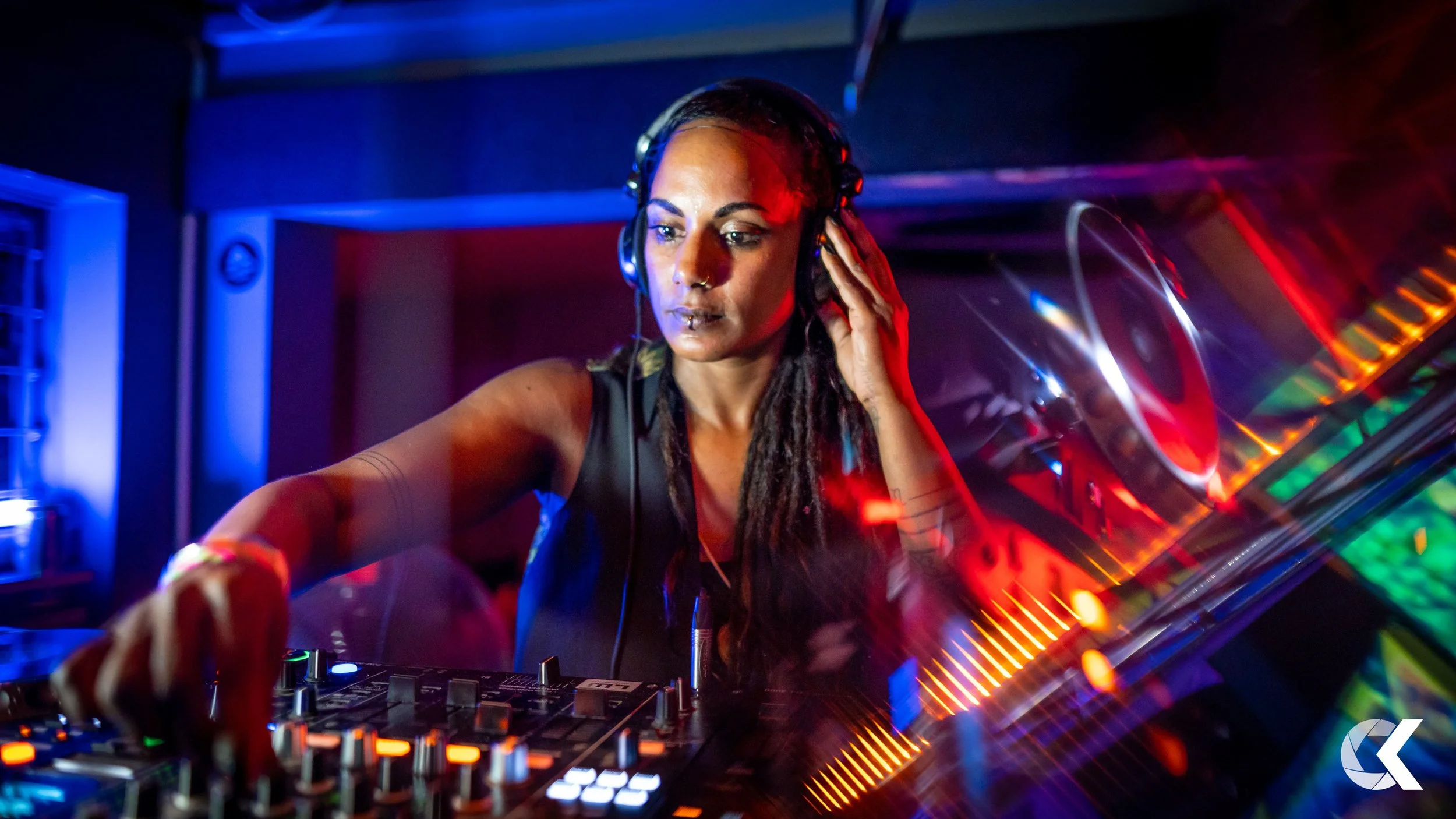 Female DJ with dreadlocks wearing headphones, operating DJ equipment in a vibrant, colorful nightclub or party setting.