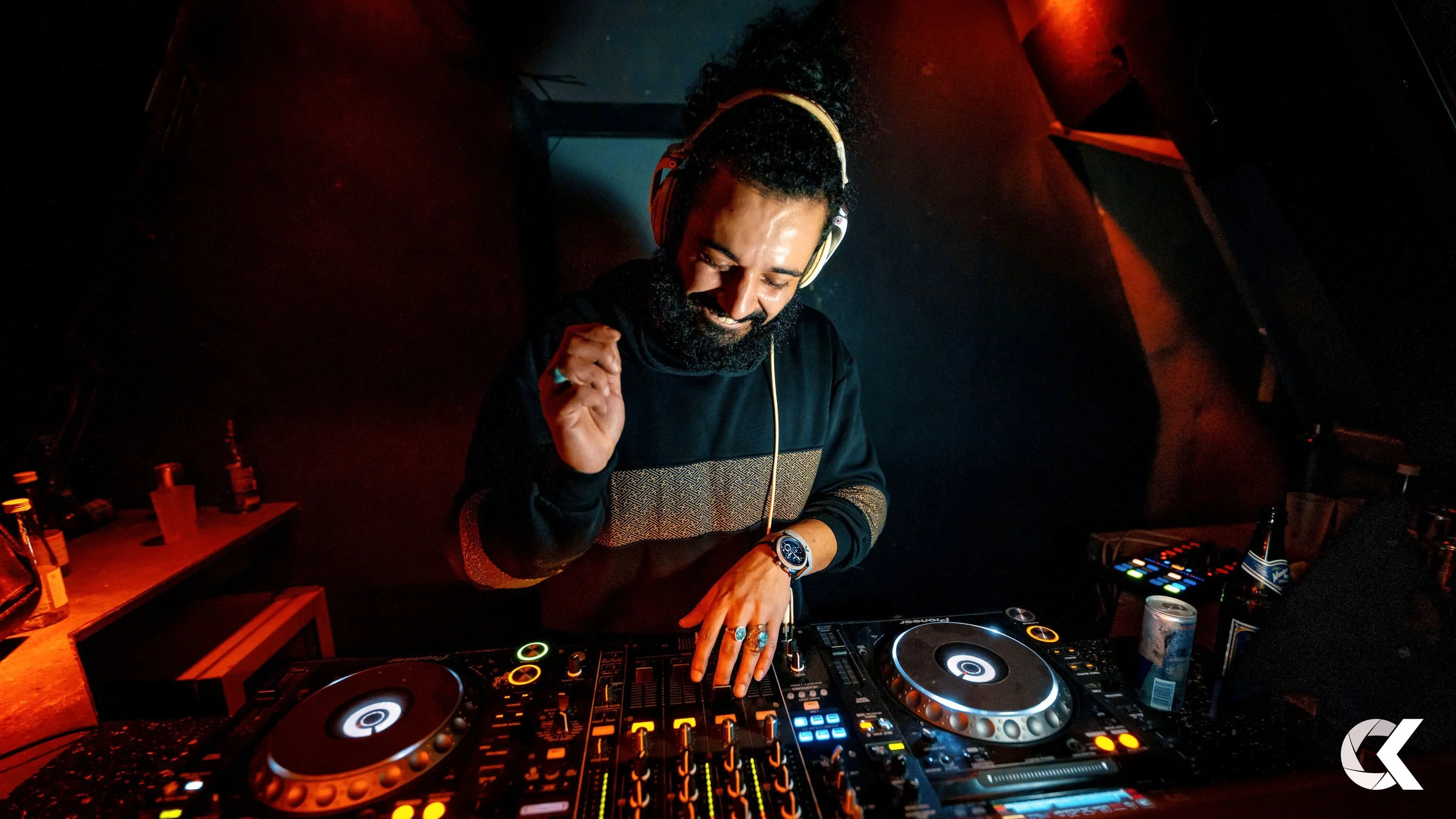 A DJ wearing headphones, glasses, and a watch, smiling and dancing behind a DJ mixing console with colorful lights in a dimly lit nightclub.