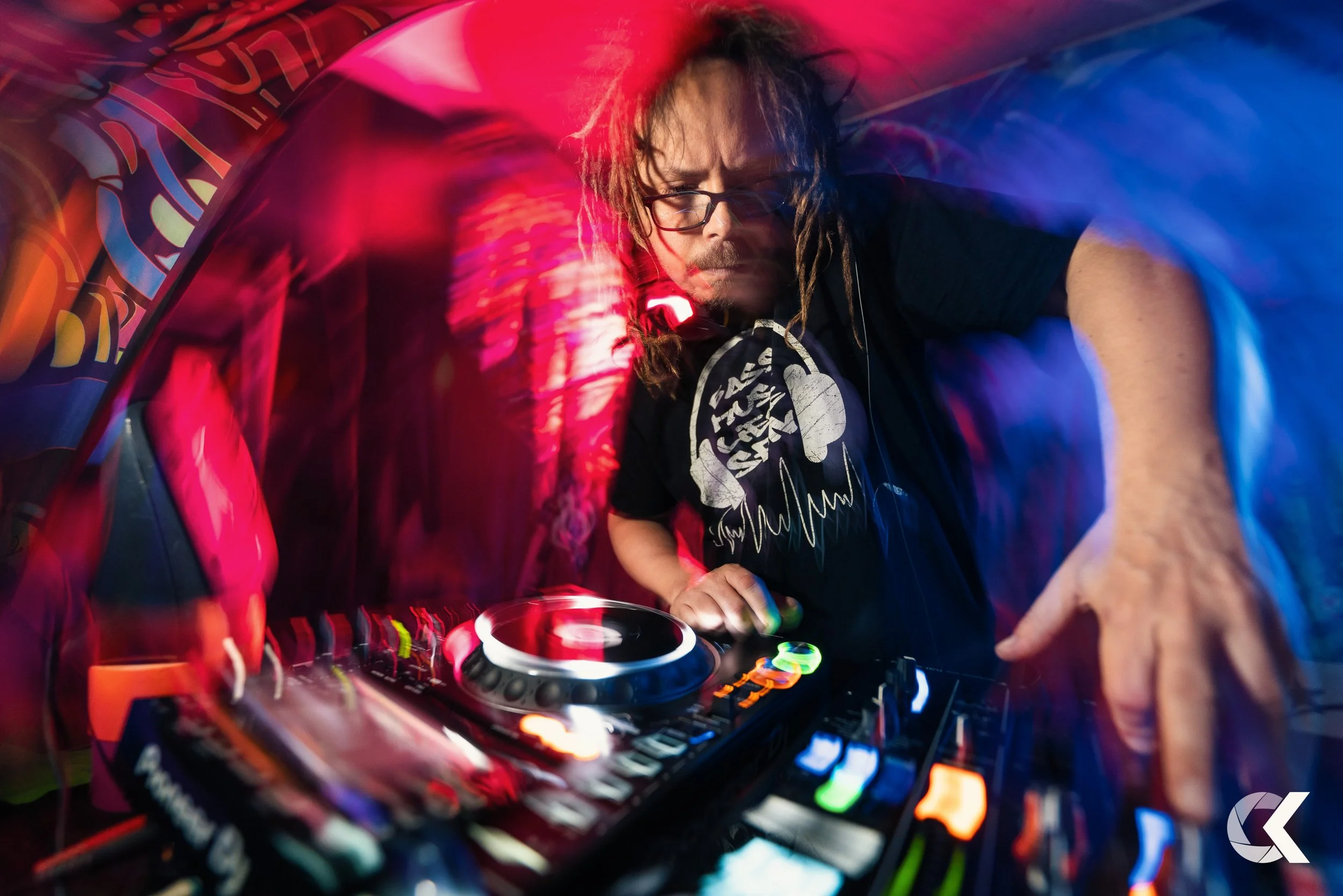 A DJ with glasses and dreadlocks is playing music at a nightclub, using a CDJ and mixer surrounded by colorful, vibrant lights.