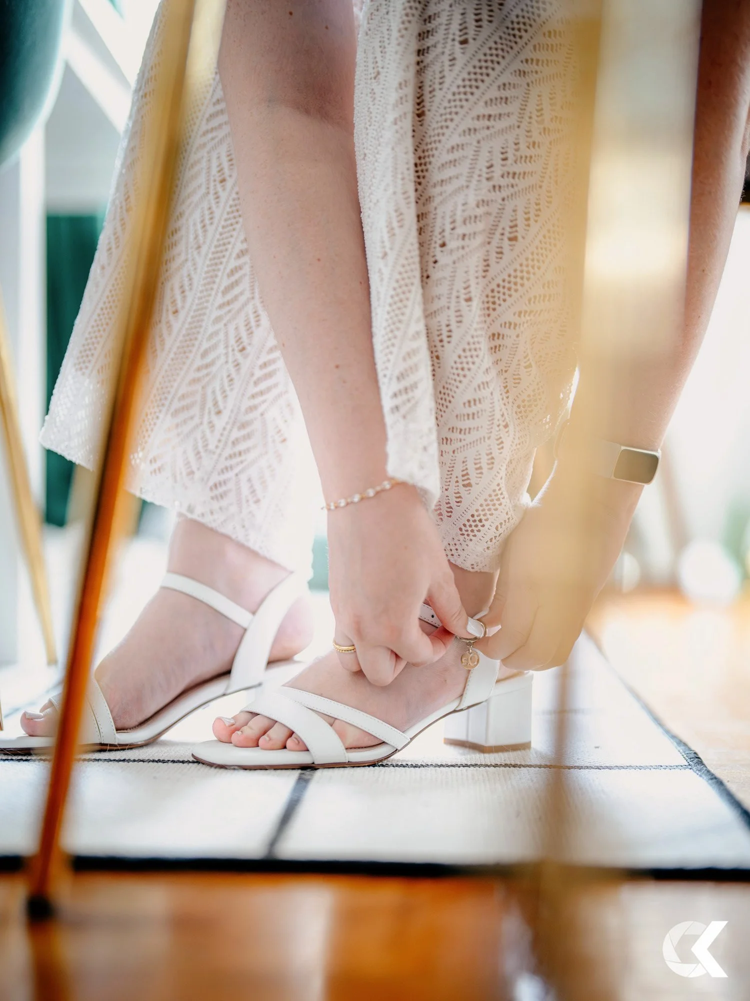 Woman putting on white heeled sandals, wearing a white lace dress, with jewelry on her wrist and fingers, standing on a woven rug.