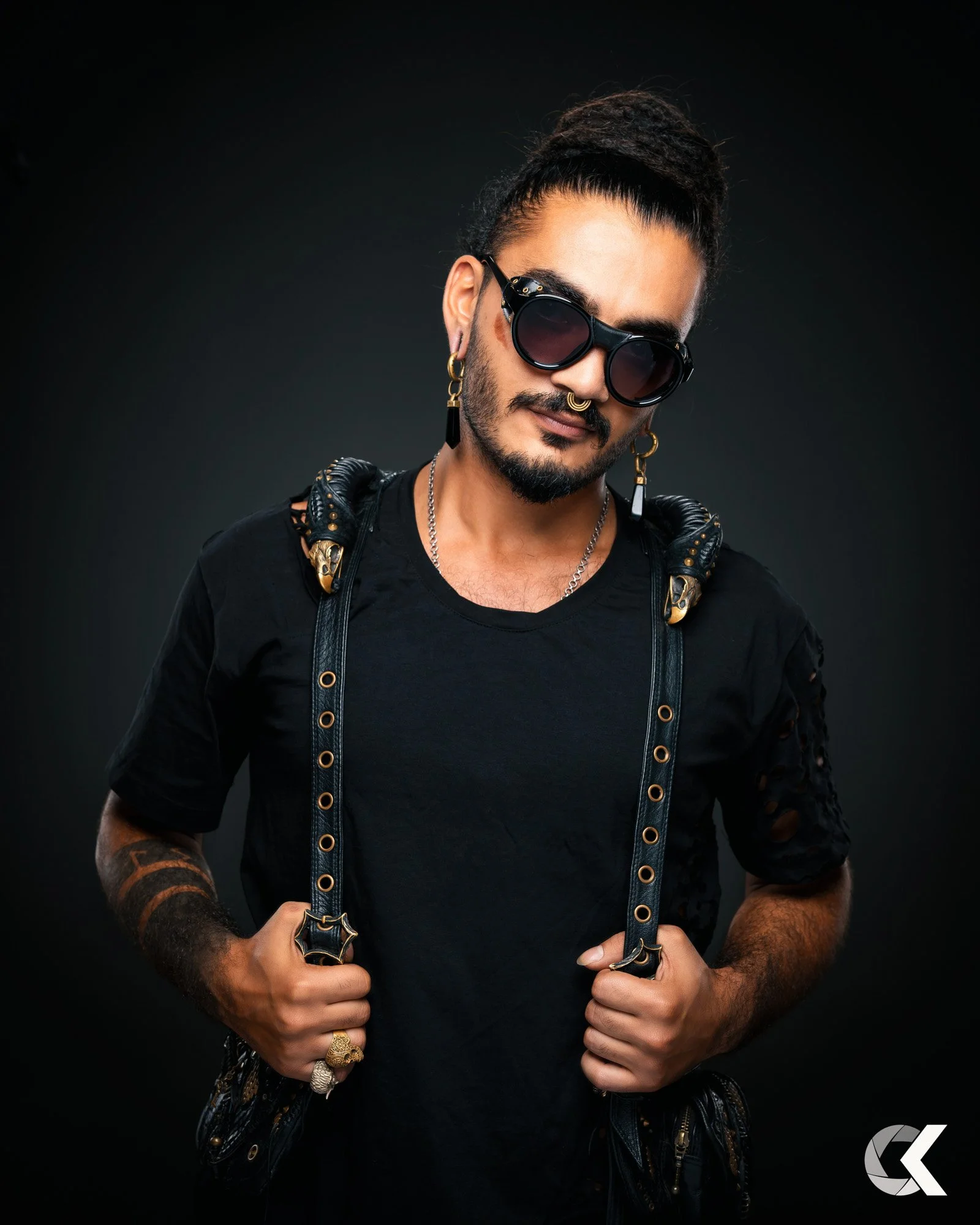 Ein stilvoller junger Mann mit dunklen Haaren, Sonnenbrille, Ohrringen, Tattoos und grünen Accessoires, der einen schwarzen Rucksack trägt, vor einem schwarzen Hintergrund.