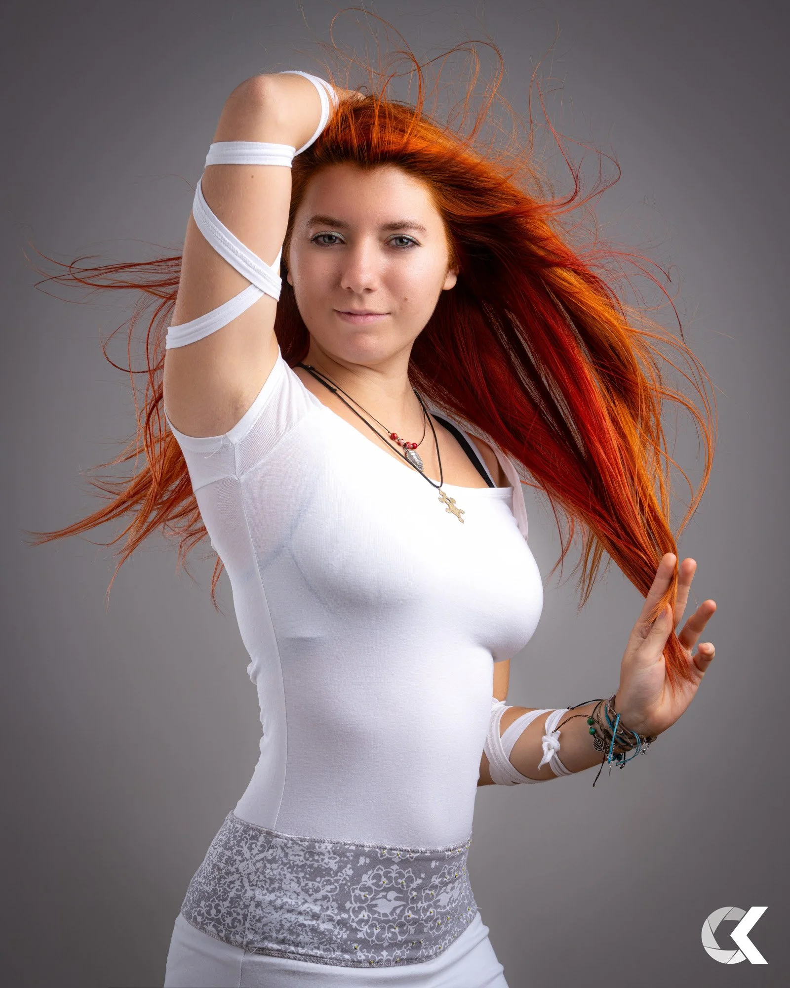 A woman with long red hair flowing, wearing a white fitted t-shirt and layered necklaces, raising one arm above her head, against a gray background.