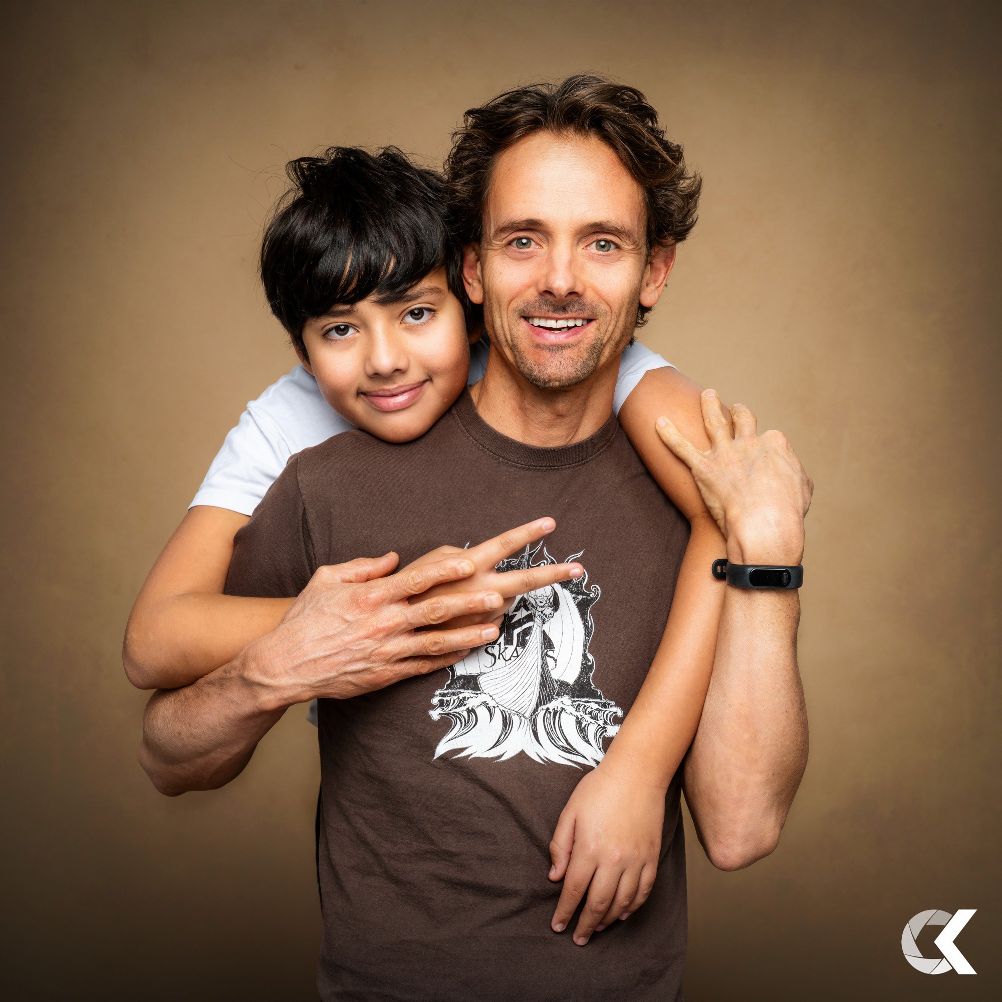 A man and a young boy smiling together, with the boy hugging the man from behind and resting his head on the man's shoulder against a plain background.