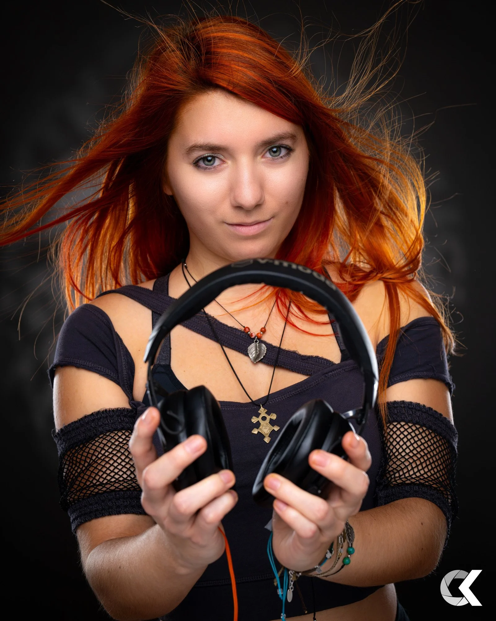 A young woman with red hair holding headphones in front of her with a serious expression, against a dark background.