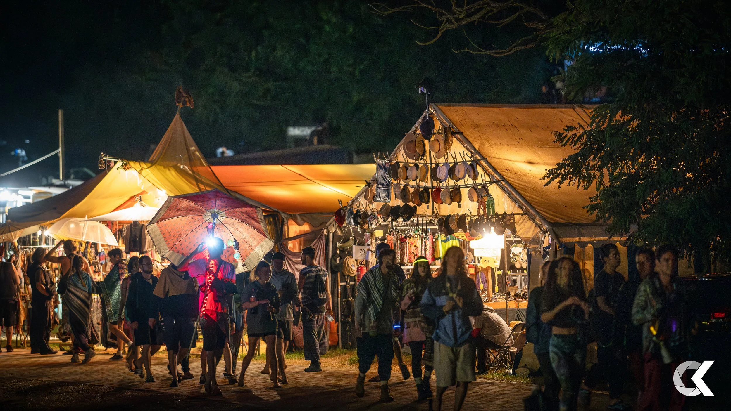 Night scene at a lively outdoor market with illuminated tents and stalls selling hats, clothing, and accessories. People walk by, some holding umbrellas, under trees in the background.