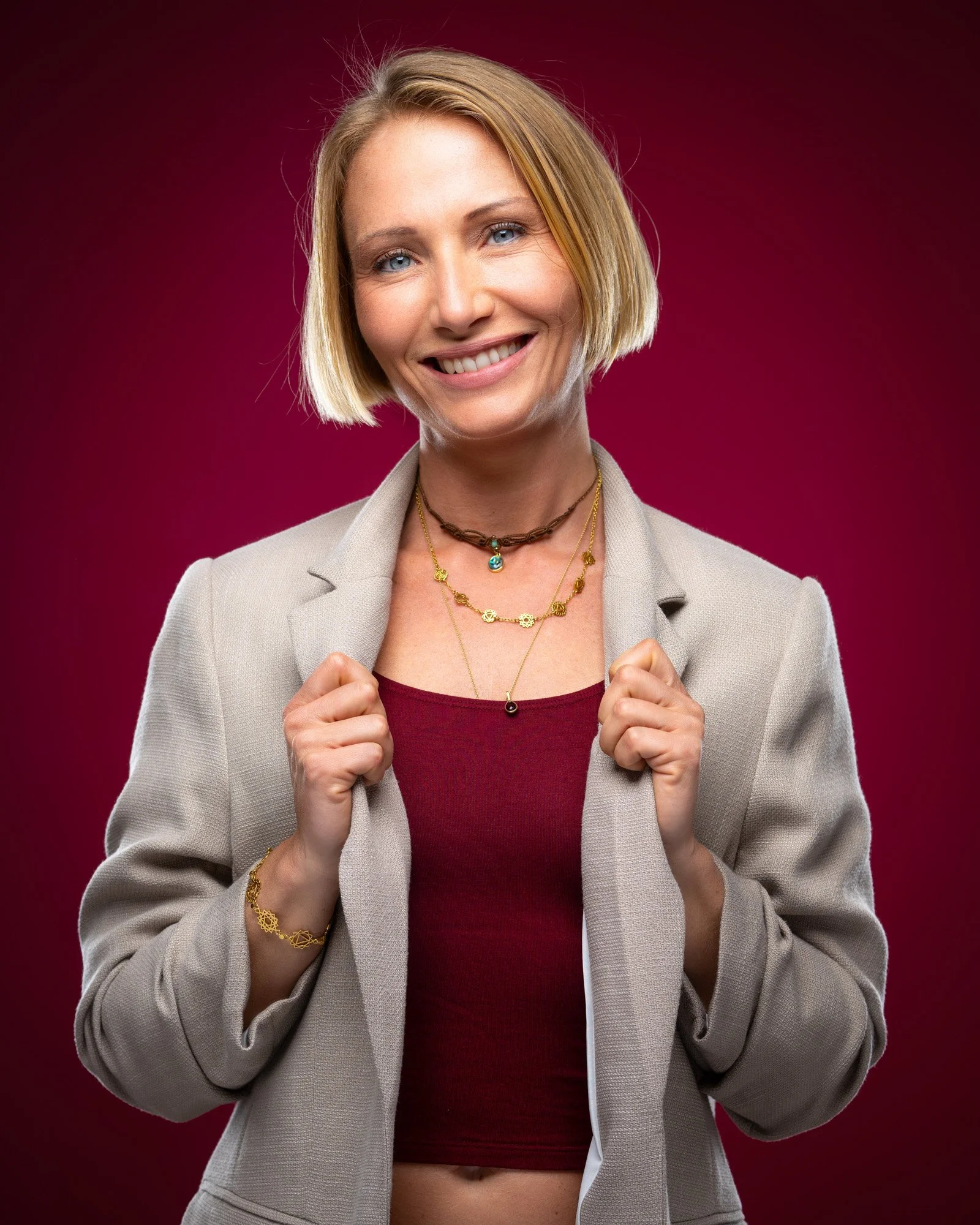 A smiling woman with short blonde hair, wearing a beige blazer over a red top, accessorized with layered necklaces, a bracelet, and a choker, standing against a dark pink background.