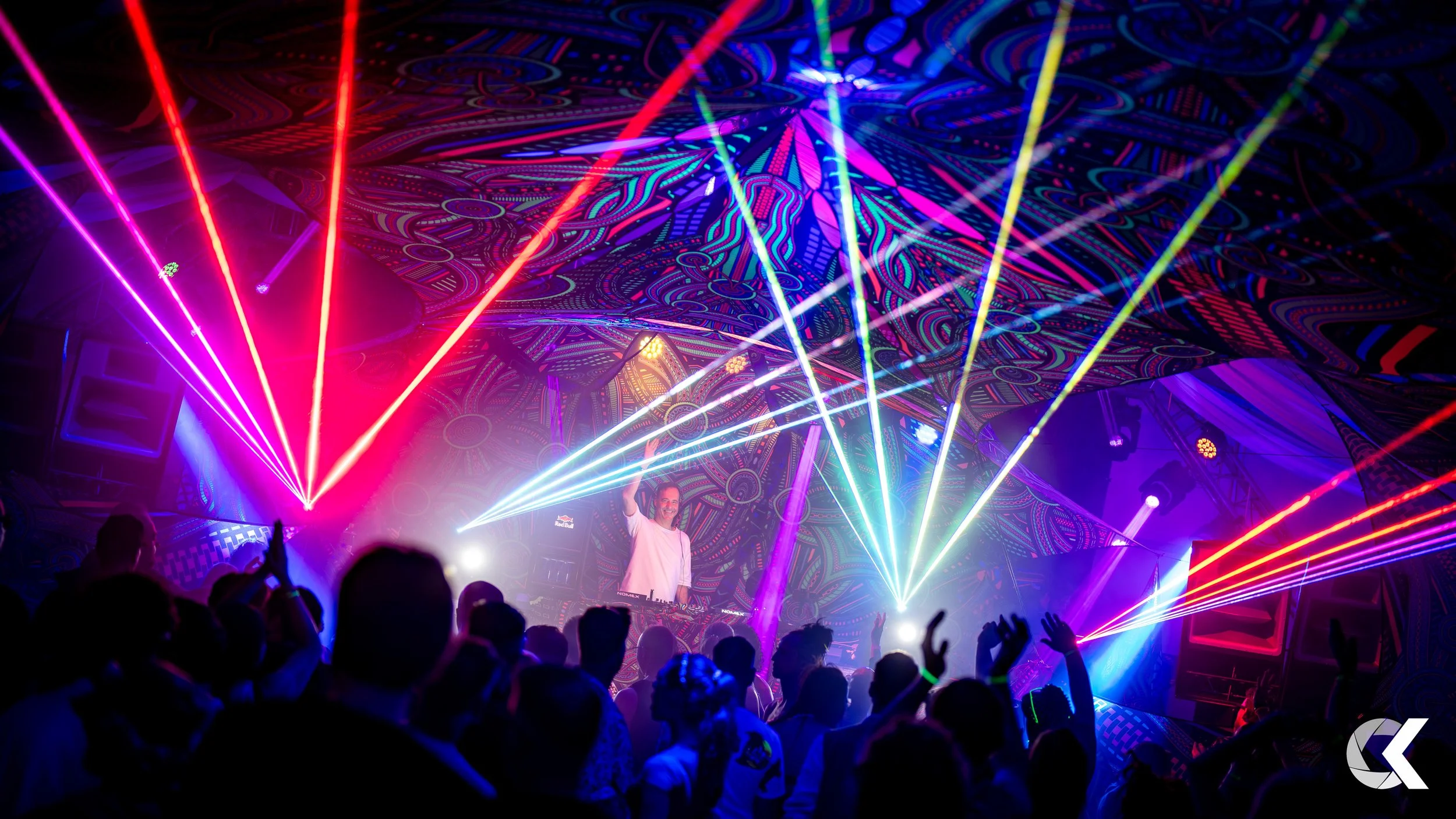 A DJ performing on stage at a nightclub with colorful laser lights and a crowd dancing.