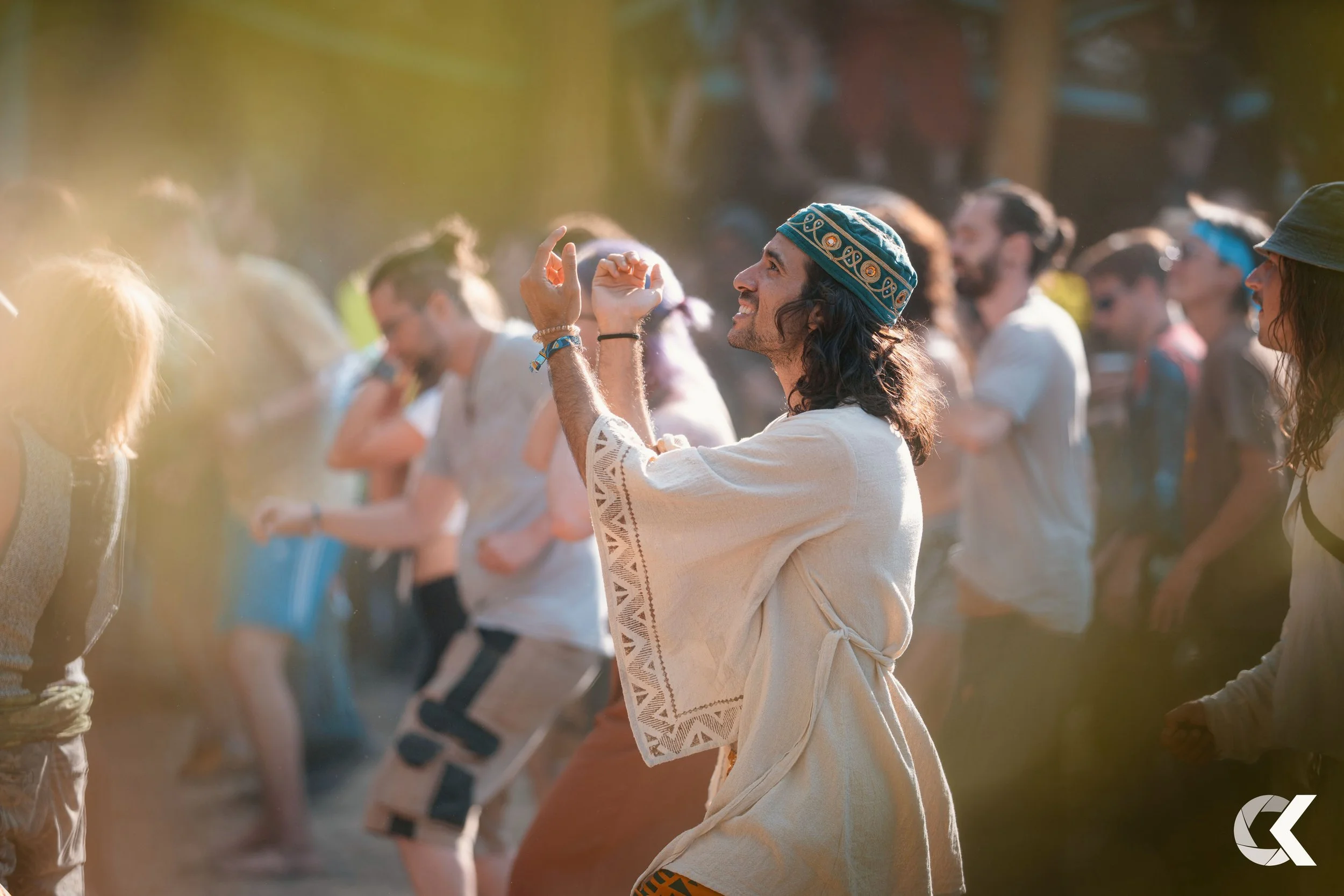 Man with long hair, wearing a tasseled headband and a cream-colored embroidered garment, smiling and dancing among a crowd at an outdoor festival.