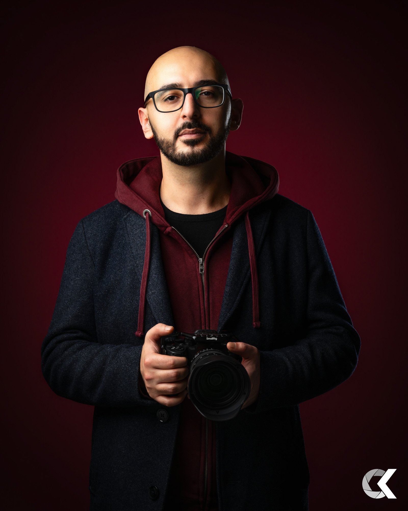 A man with glasses, a beard, and a mustache holding a camera, wearing a dark jacket over a maroon hoodie, standing against a dark red to black gradient background.