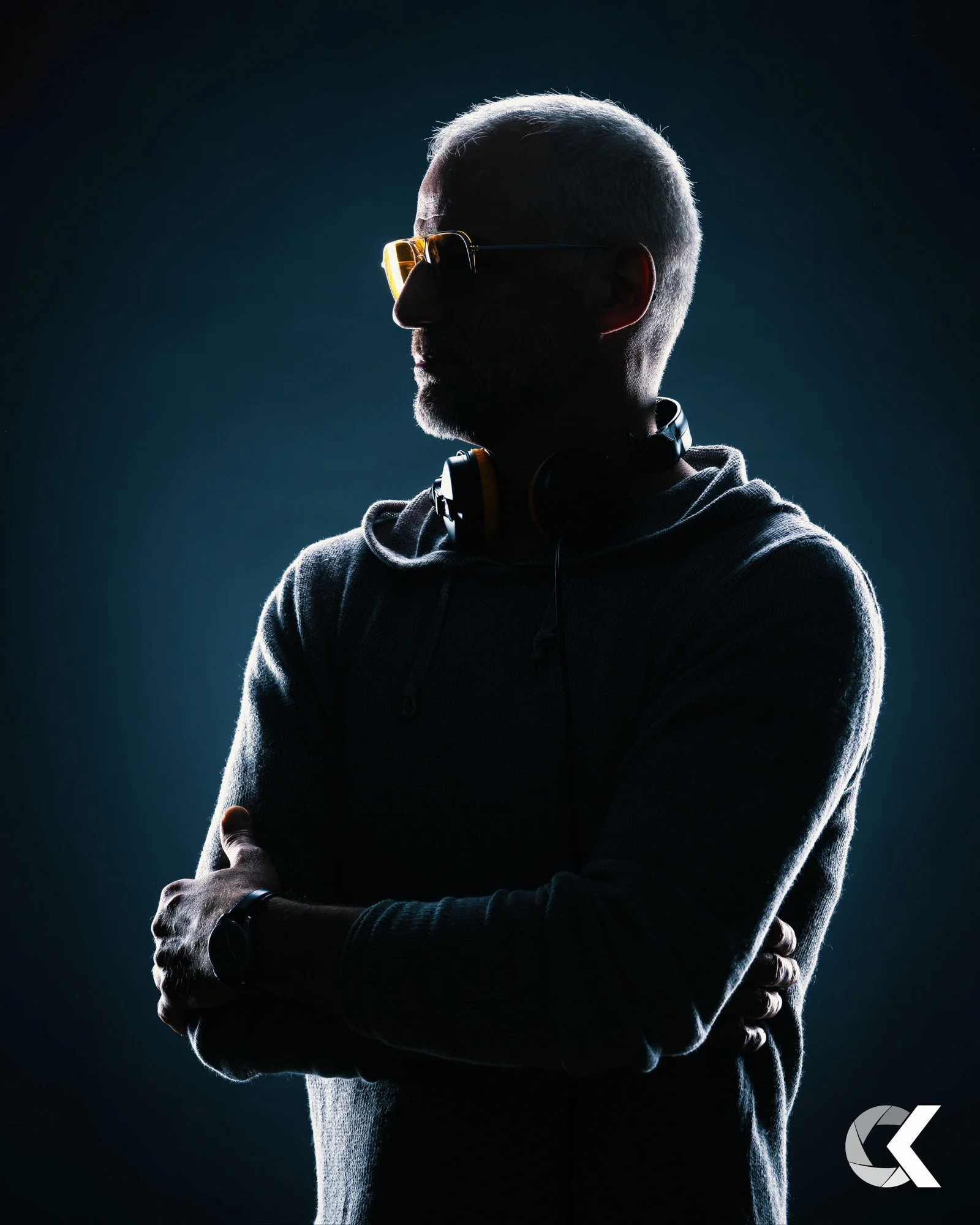 Ein osteuropäischer Mann mit Glatze, Sonnenbrille, grauem Hoodie, Kopfhörern um den Hals und einer Armbanduhr, steht vor dunklem Hintergrund, beleuchtet von seitlichem Licht.