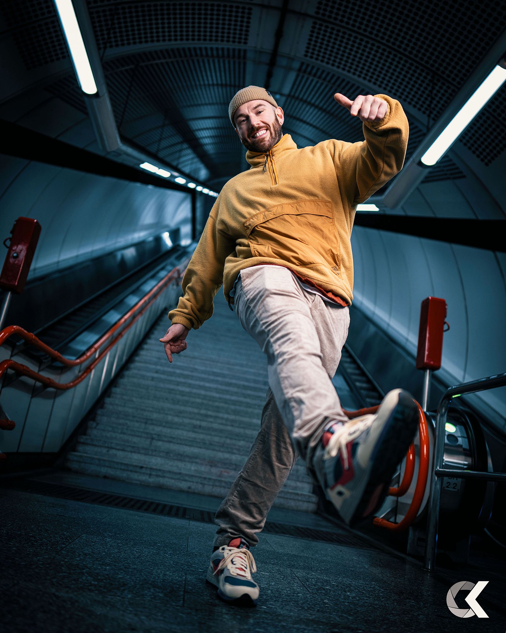 A young man in a yellow jacket, beige pants, and sneakers is smiling and balancing on one foot at the bottom of an underground escalator.