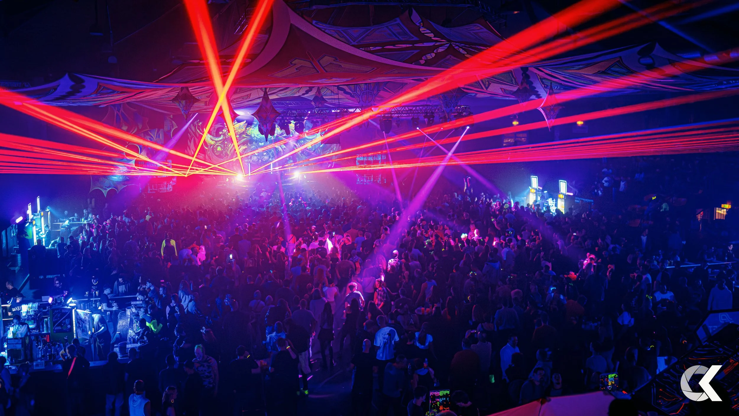A crowded concert or dance event with vibrant red, purple, and blue laser lights illuminating the large audience and stage.
