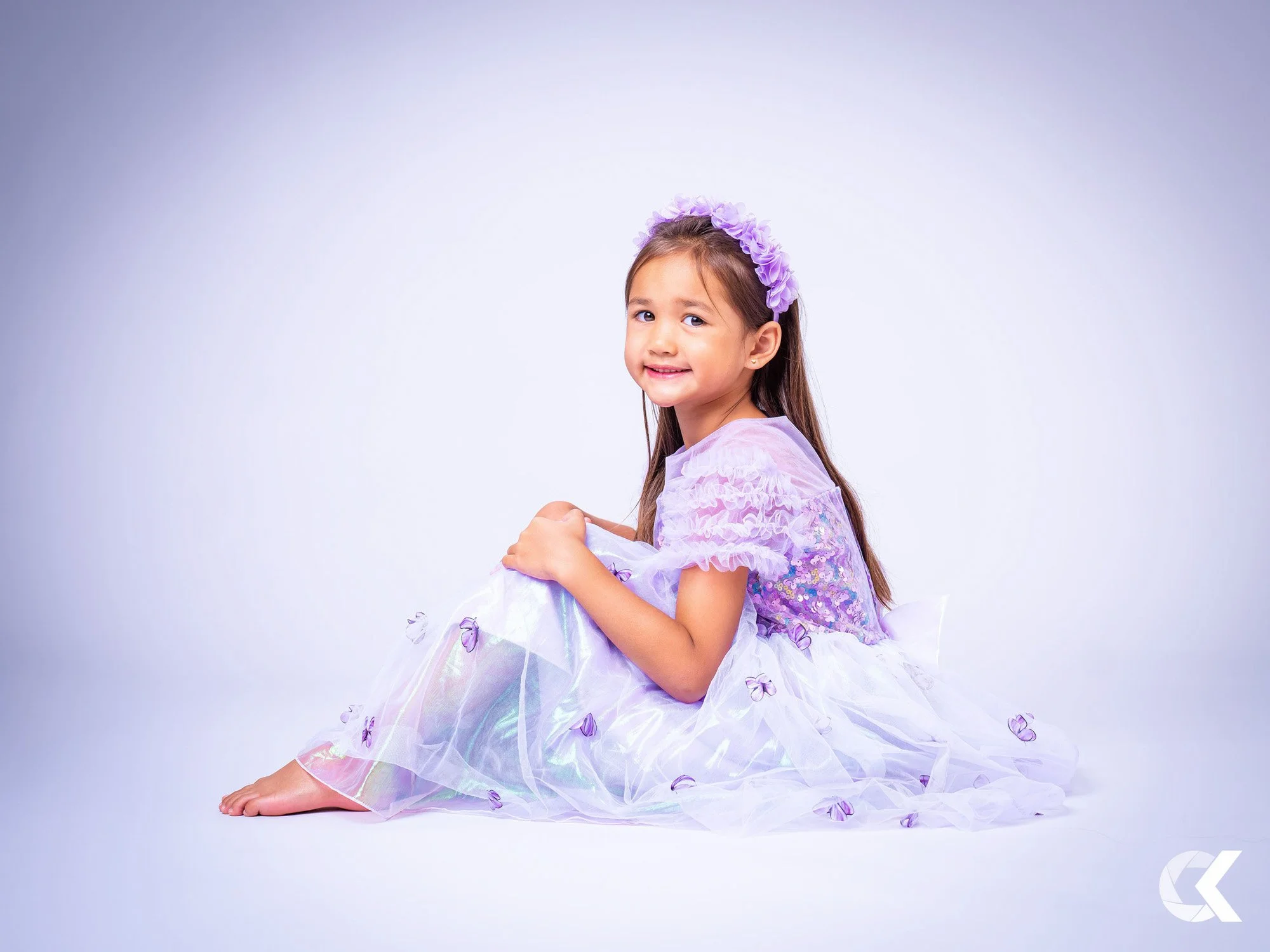 Young girl sitting on the floor, wearing a light purple fairy costume with a matching flower headband, smiling at the camera against a plain white background.